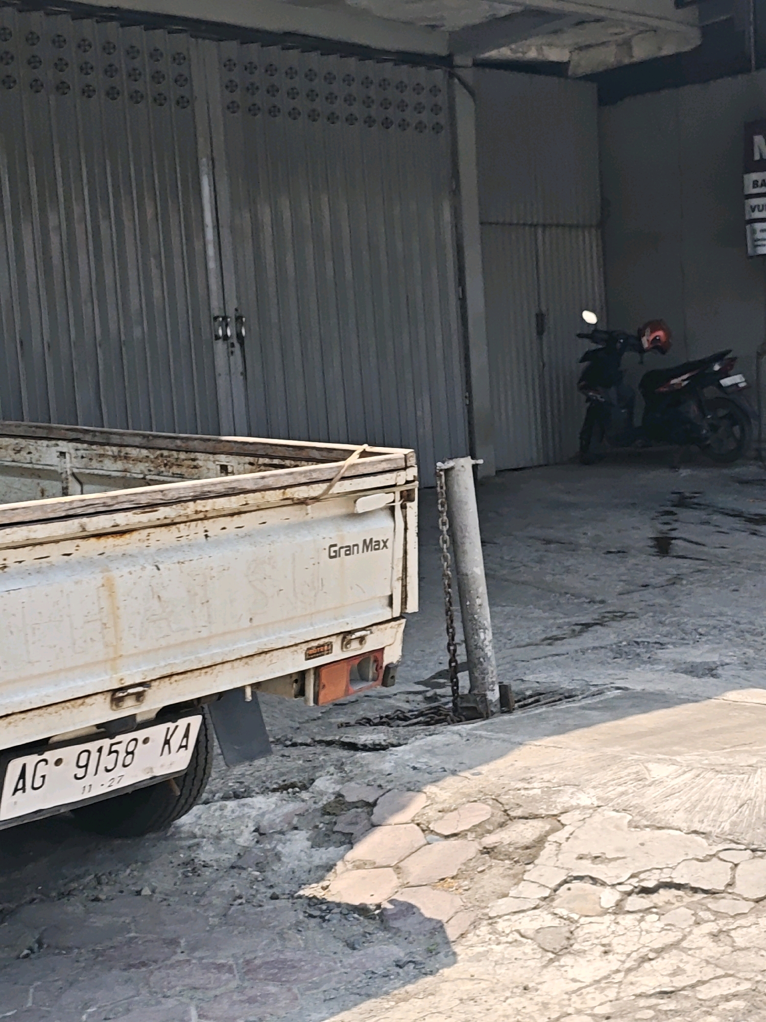 The image captures a close-up view of the rear of a white truck parked in an outdoor, urban setting that appears to be within a commercial area. The truck is slightly aged, with visible signs of wear and rust on its metal body and wooden bed frame. A license plate, reading "AG · 9158 · KA" and dated "11-27", is clearly visible. Next to the truck, a concrete post with a hanging chain stands on a cracked pavement. In the background, a corrugated metal gate is partially visible, along with two parked motorcycles, one with a red helmet on its seat. The overall scene suggests a daytime setting, with sunlight casting shadows on the ground. The context points to a location in Kediri City, Indonesia, possibly a parking lot or a service area.
