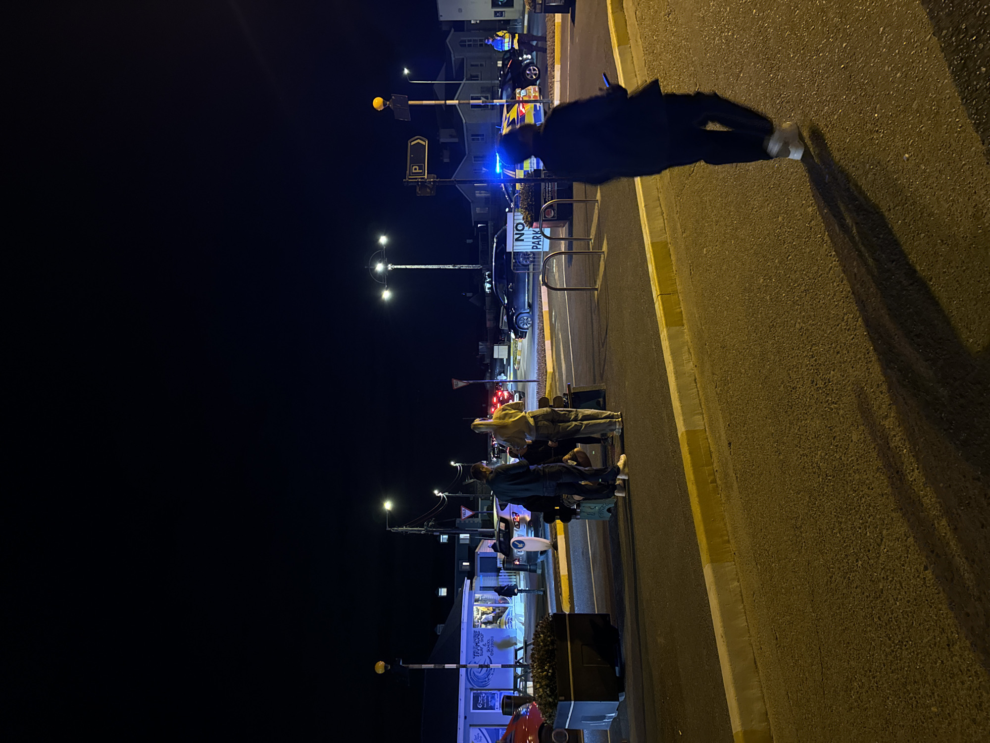 The image depicts a night scene in Tramore, Ireland, with a focus on a public area that appears to be near a street or parking lot. In the foreground, a person walks away from the camera, casting a long shadow on the asphalt. The environment is illuminated by streetlights and the blue and yellow flashing lights of what appear to be police vehicles, suggesting a potential incident or ongoing operation.
Several individuals are visible in the mid-ground, some standing and others sitting on a bench. They are gathered near a shop labeled "TRAMORE SURF & SHOP." Luggage is present, indicating travel or a waiting situation. The shop's interior is visible and lit, showing some activity within.
In the background, houses and other buildings are dimly lit. The overall atmosphere is dark, typical of a nighttime setting. The presence of police vehicles and their flashing lights is a prominent feature of the scene. There is a "NO PARKING" sign visible, further contextualizing the area. The weather appears clear, with no signs of rain or extreme conditions.