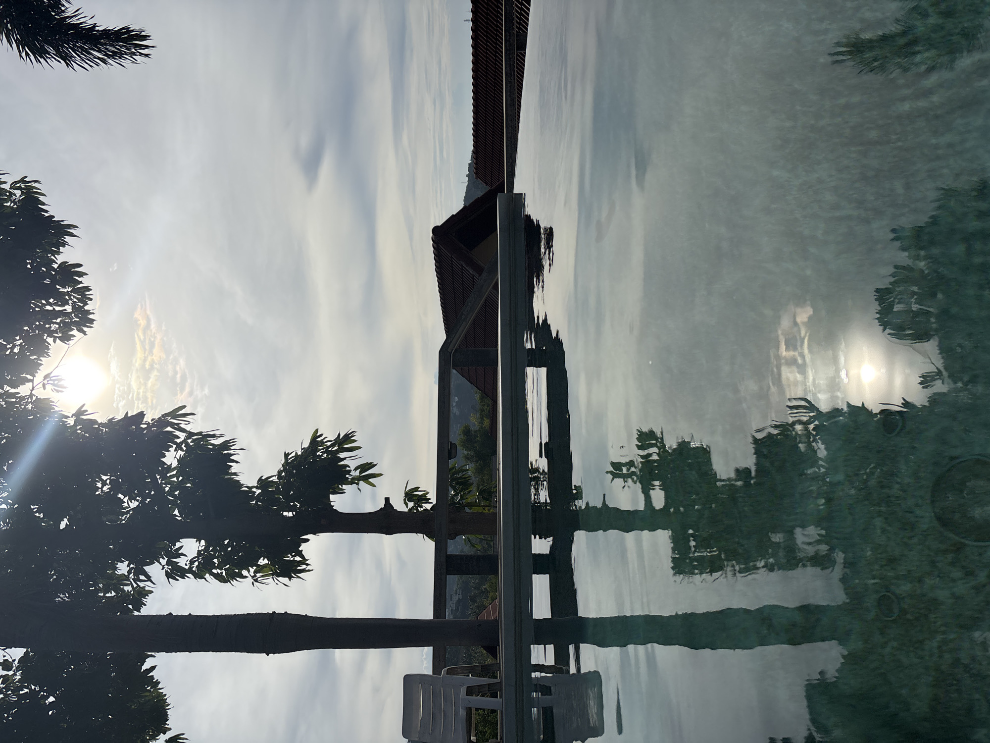 The image shows a serene view of an infinity pool at Ban Bang Rak, Thailand, during a bright, overcast day. The pool's surface reflects the sky, trees, and the surrounding architecture. In the foreground, the clear, tranquil water of the pool occupies the majority of the frame. The pool's edge, a light-colored ledge, is visible, and beyond it, a wooden railing separates the pool area from a slight drop.
On the deck, a single white plastic chair stands to the left, positioned near two silhouetted trees. One tree is notably taller and thinner with a textured trunk, while the other is shorter and bushier, with a canopy of leaves. The foliage of these trees, along with a palm frond visible in the upper right corner, casts shadows and is reflected in the water.
Behind the railing, the landscape unfolds with the distinctive reddish-brown tiled roofs of houses nestled amongst lush green vegetation. The sky is a mix of bright white clouds and areas of pale blue, with the sun's glare creating a strong lens flare effect in the upper center of the image. The overall atmosphere is peaceful and invites relaxation, capturing the essence of a tropical getaway.