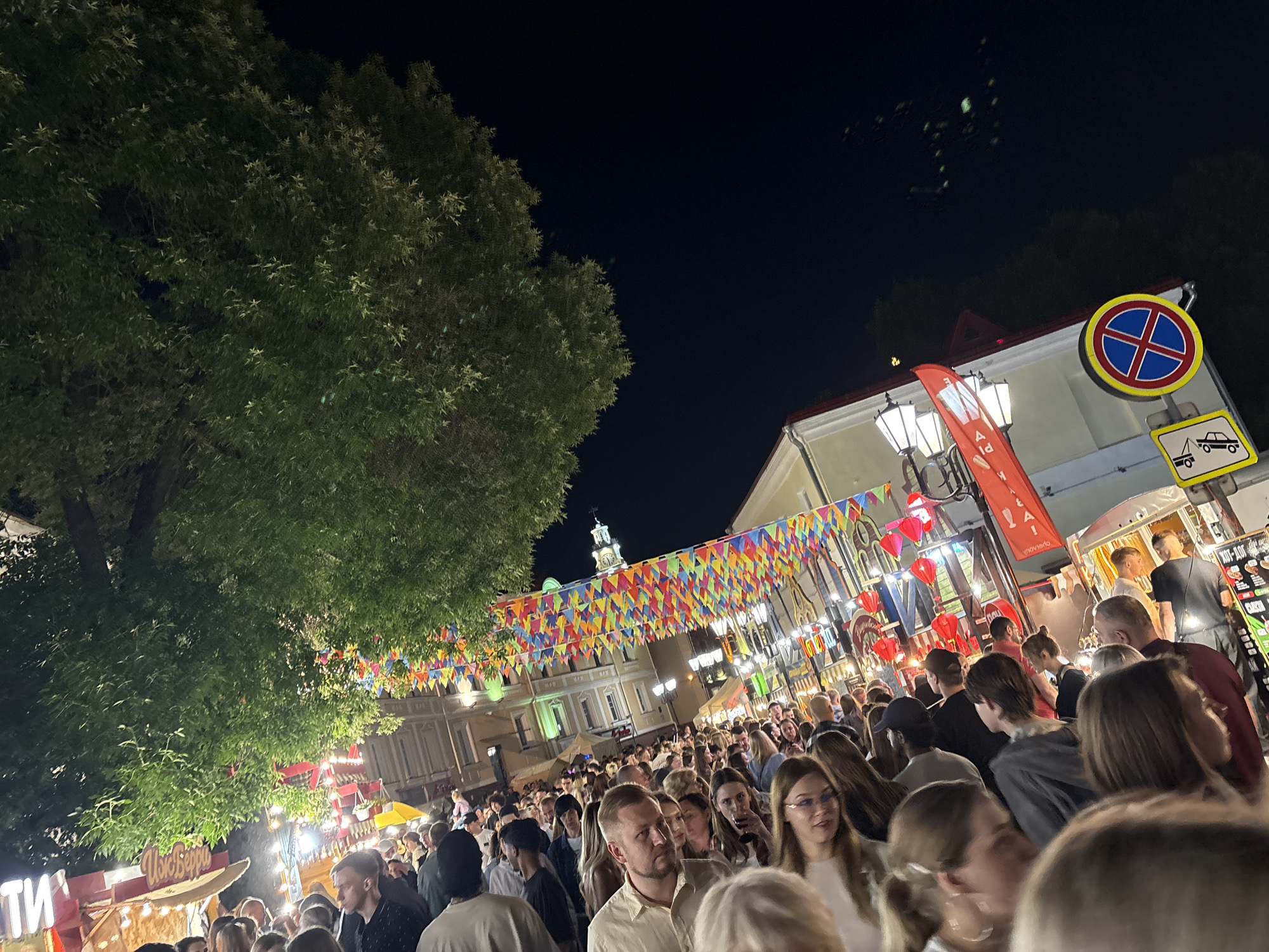 The image depicts a crowded outdoor night market or festival, likely in Vitebsk, Belarus, given the Cyrillic text visible on a banner. A large, leafy tree dominates the left side of the frame, its branches illuminated by artificial lights. Overhead, colorful triangular flags are strung across the street, creating a festive atmosphere. A building with white and cream-colored walls and a red-tiled roof is visible in the background, adorned with street lamps and red lanterns. Traffic signs, including a no-stopping sign and a towing warning, are mounted on a pole near the building. A dense crowd of people, dressed in casual attire, are the central focus of the scene. They are looking in various directions, some engaged in conversation, suggesting a lively and bustling event. Stalls with visible signage, some featuring Cyrillic script, are set up, indicating food vendors or merchandise. The overall impression is that of a vibrant community gathering at night.