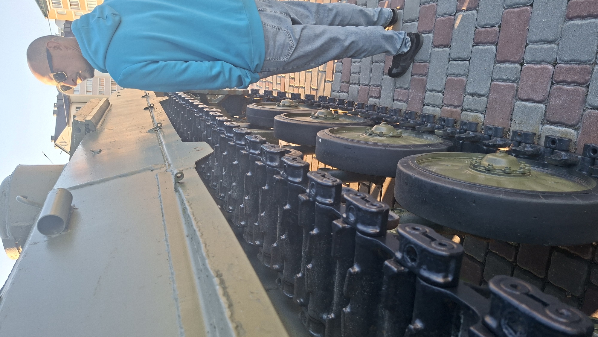 A man wearing sunglasses, a light blue hoodie, and light gray jeans stands on a brick pathway next to a large, olive-green tracked vehicle, possibly a tank. The vehicle's tracks and several road wheels are visible. Buildings are in the background. The scene appears to be outdoors during daylight.