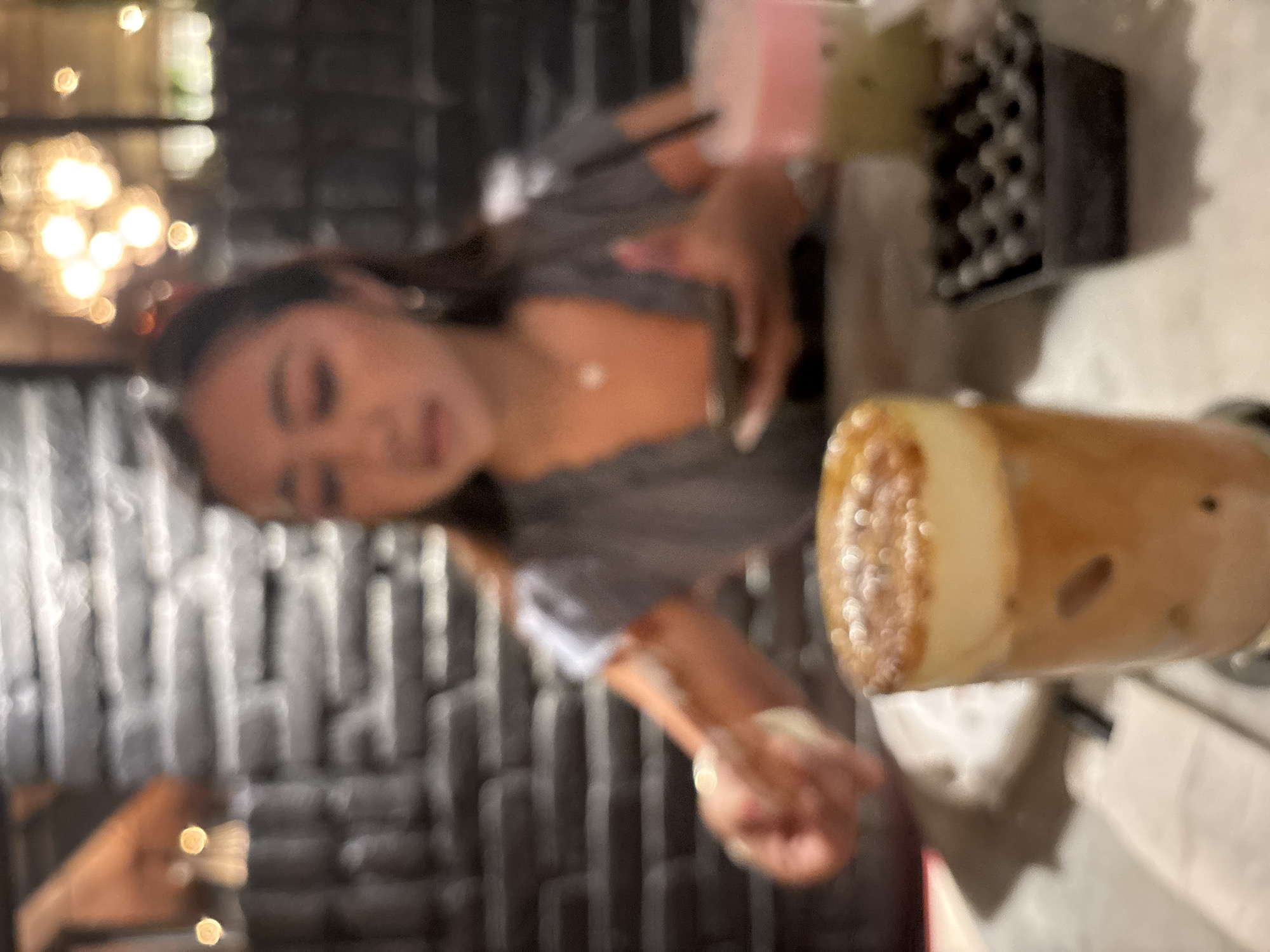 The image depicts a blurry, indoor scene. A woman is seated at a table, holding a mobile phone in her right hand and an object that appears to be a cigarette or a rolled paper in her left. She is wearing a grey top and a necklace. In front of her is a glass with a layered drink topped with foam and a sprinkle of brown sugar. To the right, another drink with pink and green layers is visible. Behind the table is a textured brick wall. The setting suggests a restaurant or cafe in North Kuta, Indonesia.