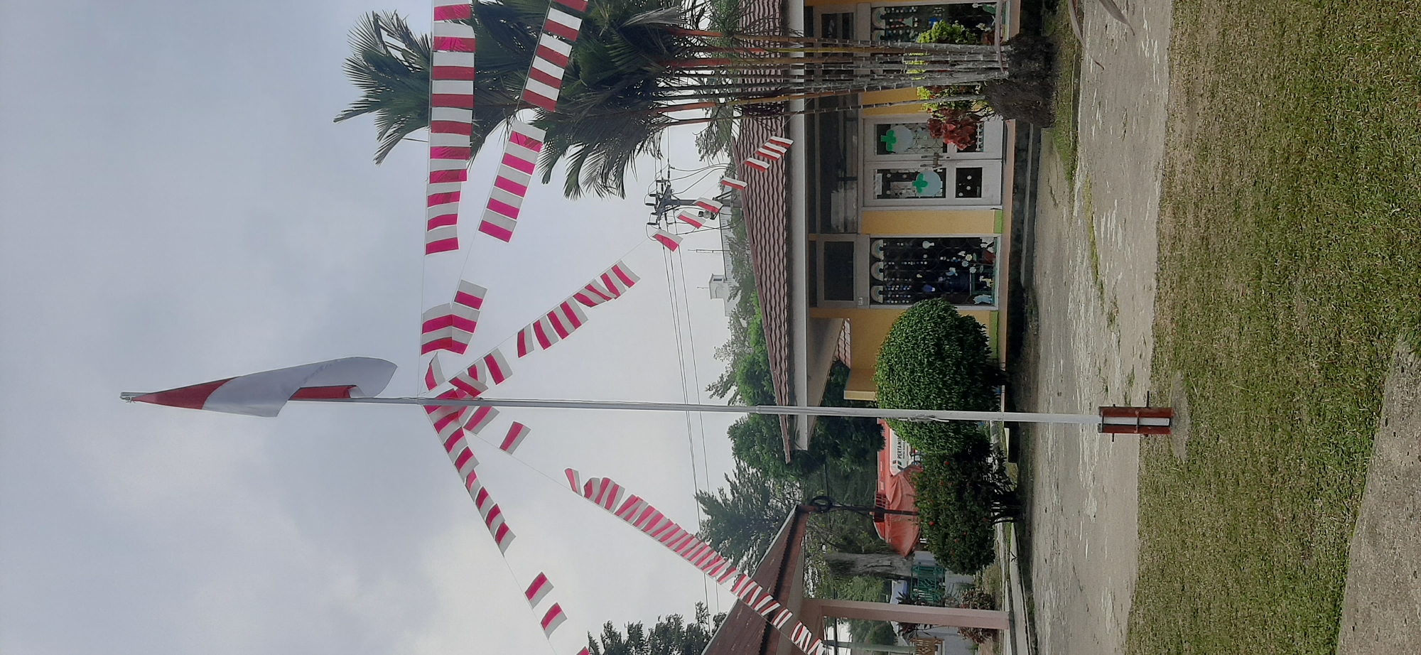 The image shows a school building in Jambi, Indonesia, decorated with Indonesian flags and red and white streamers. The building is painted yellow with a brown tiled roof. There are trees and bushes in front of the building, and a flagpole with the Indonesian flag is visible. The sky is cloudy. A large red vehicle, possibly a fire truck, is parked in the background. There are no people visible in the image.