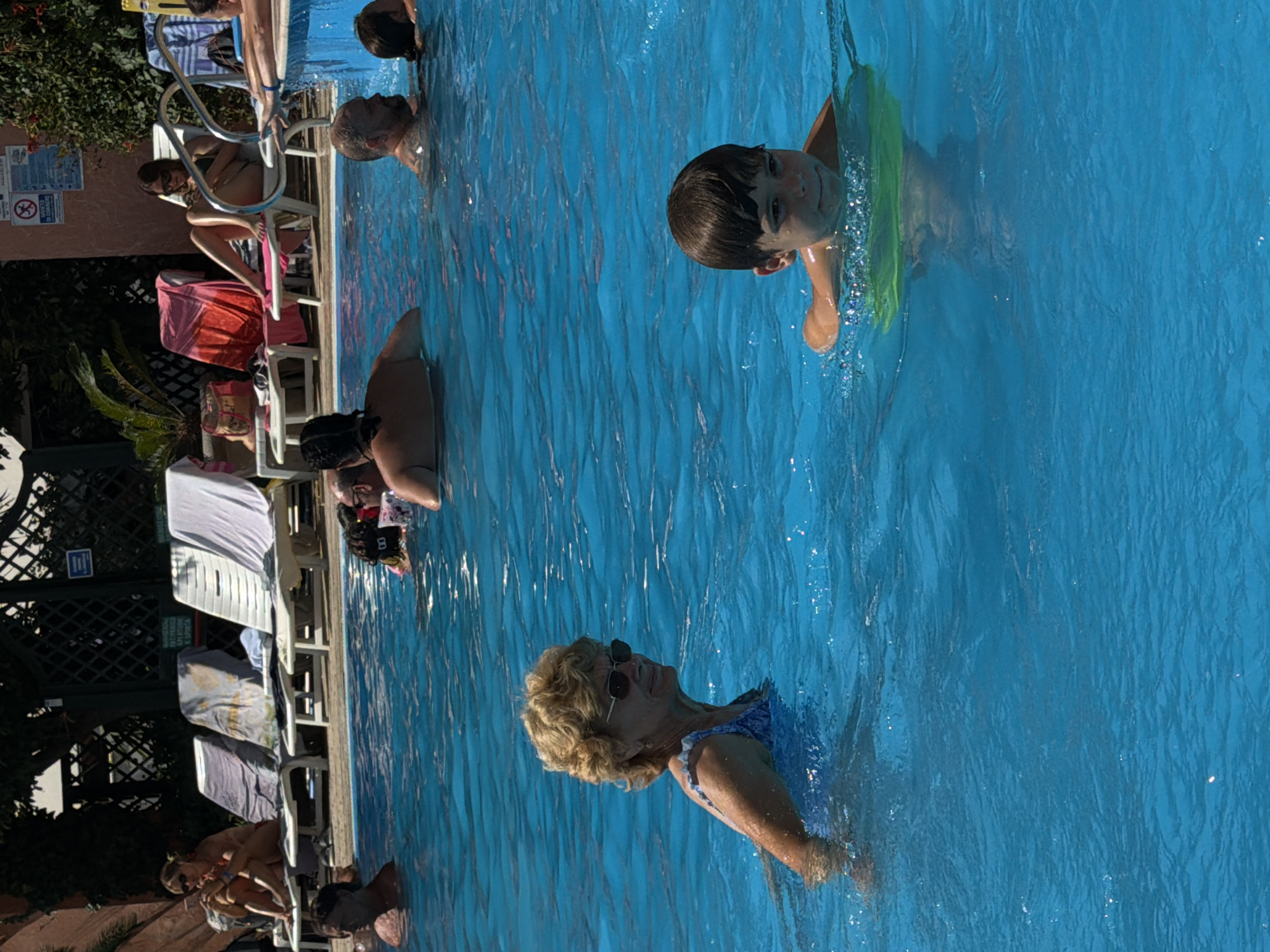 In this high-angle shot, a group of people enjoy a sunny day at an outdoor swimming pool. The bright blue water fills most of the frame, with some of the pool deck and lounge chairs visible on the left side. The scene appears to be in Gatteo, Italy, suggesting a warm, pleasant atmosphere.
Several individuals are in the pool. In the foreground, an older woman with blonde, permed hair and sunglasses is swimming, looking up and to her right. She is wearing a blue swimsuit. Further back, a young boy with dark hair is swimming, wearing a green floatation device around his waist. He is looking towards the camera with a slight smile. Other swimmers and people relaxing on lounge chairs are also visible.
On the left side of the frame, several white plastic lounge chairs are lined up, some occupied by people. Towels are draped over some of the chairs. Behind the chairs, a dark green fence and lush green foliage can be seen. A small, white sign with blue text is affixed to the fence. The lighting suggests it is daytime, likely mid-morning or afternoon, with the sun casting bright light on the water. The overall mood is one of leisure and recreation.