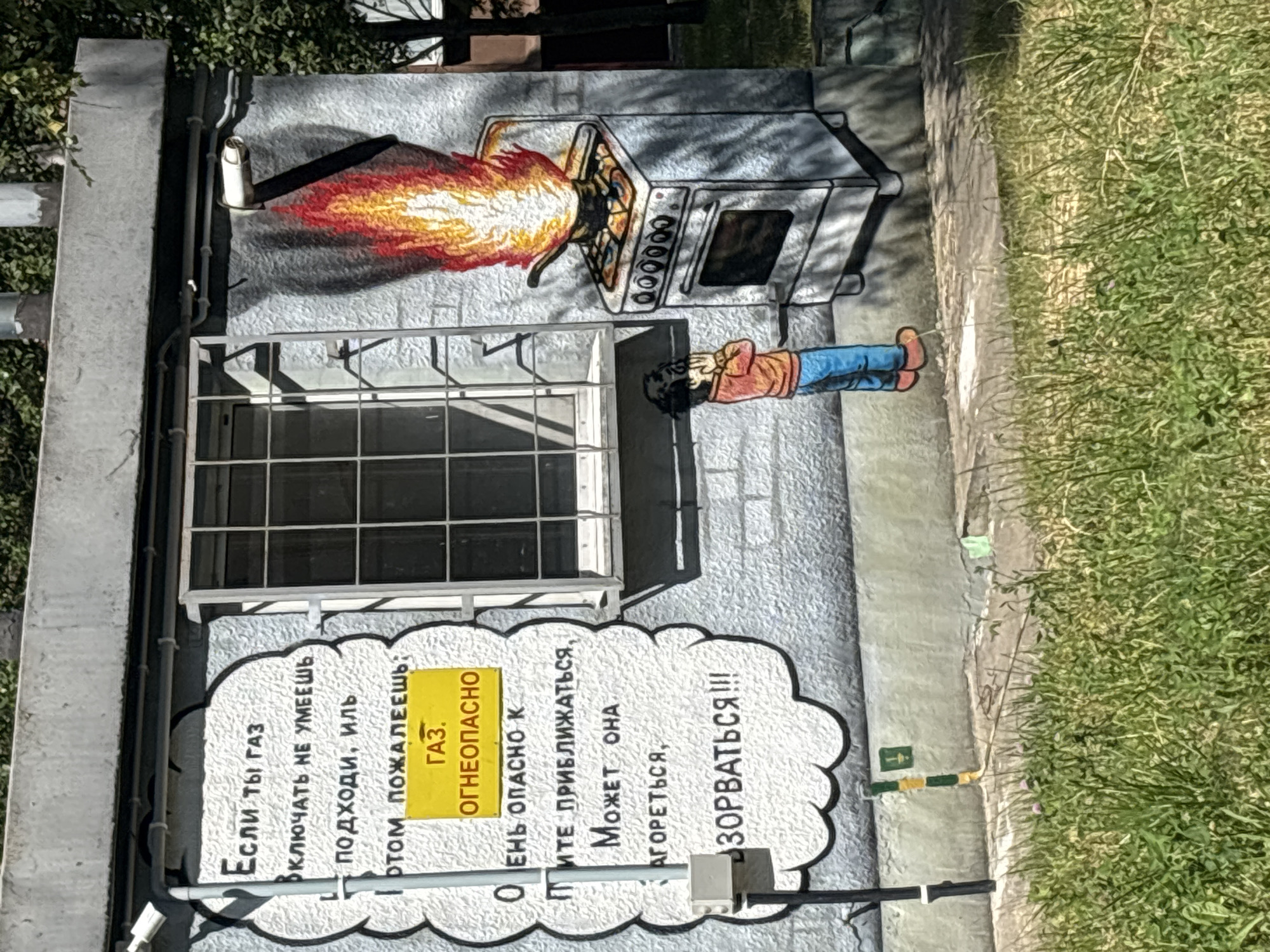 This is a photograph of a mural painted on the exterior of a building, likely in an urban setting given the concrete wall and visible piping. The mural depicts a cartoonish scene with a gas stove spewing a large flame, and a person lying on the ground covering their face with their arms. The style is illustrative and somewhat humorous, though the subject matter is a warning.
The mural includes several lines of text in Russian. On a yellow rectangular sign, it reads "ГАЗ. ОГНЕОПАСНО" which translates to "GAS. DANGEROUSLY FLAMMABLE". A larger speech bubble emanating from the scene contains more text, including "Если ты газ ВКЛЮЧАТЬ НЕ УМЕЕШЬ ПОДХОДИ, ИЛЬ ПОТОМ ПОЖАЛЕЕШЬ" which means "If you are gas, and don't know how to turn it on, come closer, or you will regret it later". Further text in the speech bubble warns "НЕ ПРИБЛИЖАТЬСЯ, МОЖЕТ ОНА АГОРЕТЬСЯ, ЗОРВАТЬСЯ!!!" translating to "DO NOT APPROACH, IT MAY CATCH FIRE, EXPLODE!!!".
The scene appears to be set outdoors during the daytime, with natural light illuminating the mural and the surrounding grassy area. The overall impression is a public safety warning depicted in a graphic and attention-grabbing artistic style.