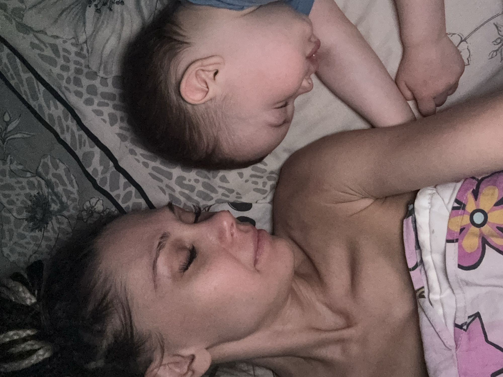 A photograph shows a woman and a child lying on a bed. The woman, facing to the left, has her eyes closed and appears to be sleeping. A patterned blanket is partially covering her torso. Above her head and at an angle, a child lies with their head tilted back, also seemingly asleep. The child's arm rests near the woman. The bedding has a patterned design. The location is not discernible from the image. There is no indication it is taken in Rusanovo, Russia.