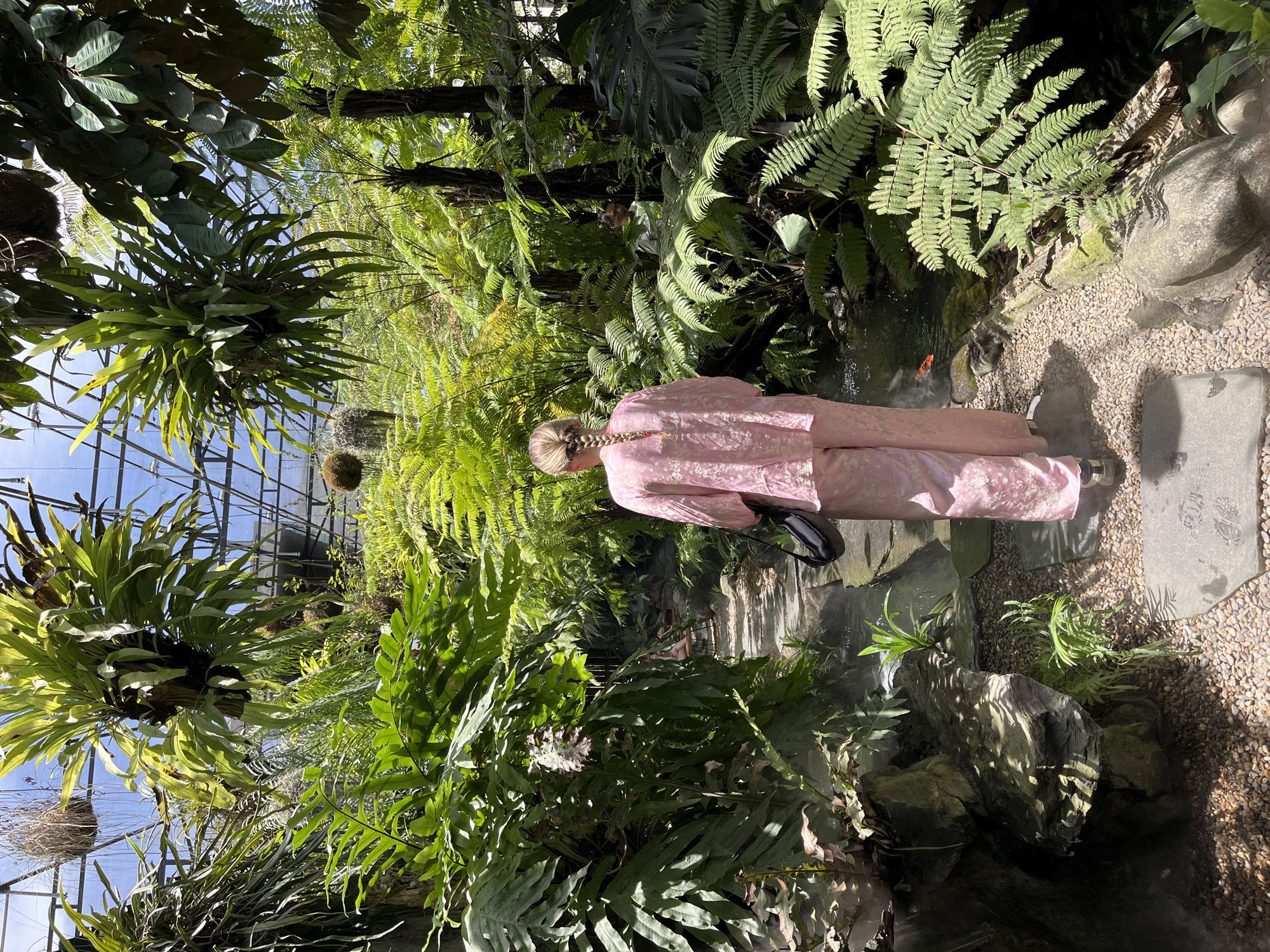 A woman with a blonde braid stands beside a small pond containing koi fish in what appears to be a lush botanical garden. She is wearing a pink, patterned pajama-style outfit and holding a black handbag. The scene is filled with various ferns and tropical plants. Above, a structure with a glass roof is visible, suggesting an indoor greenhouse or conservatory. The location is Đà Nẵng, Vietnam.