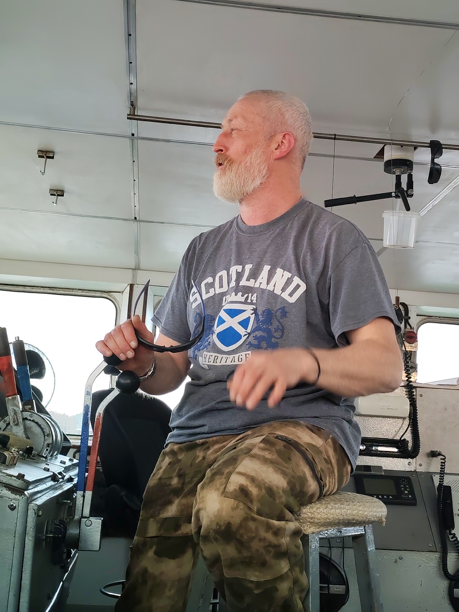 A man with a gray beard and a t-shirt that says "Scotland" is at the helm of a vessel. He is wearing camouflage pants and sitting on a cushioned stool. The interior of the vessel is utilitarian, with metal paneling and various controls and communication devices. The image was taken in Anisimovo, Russia.