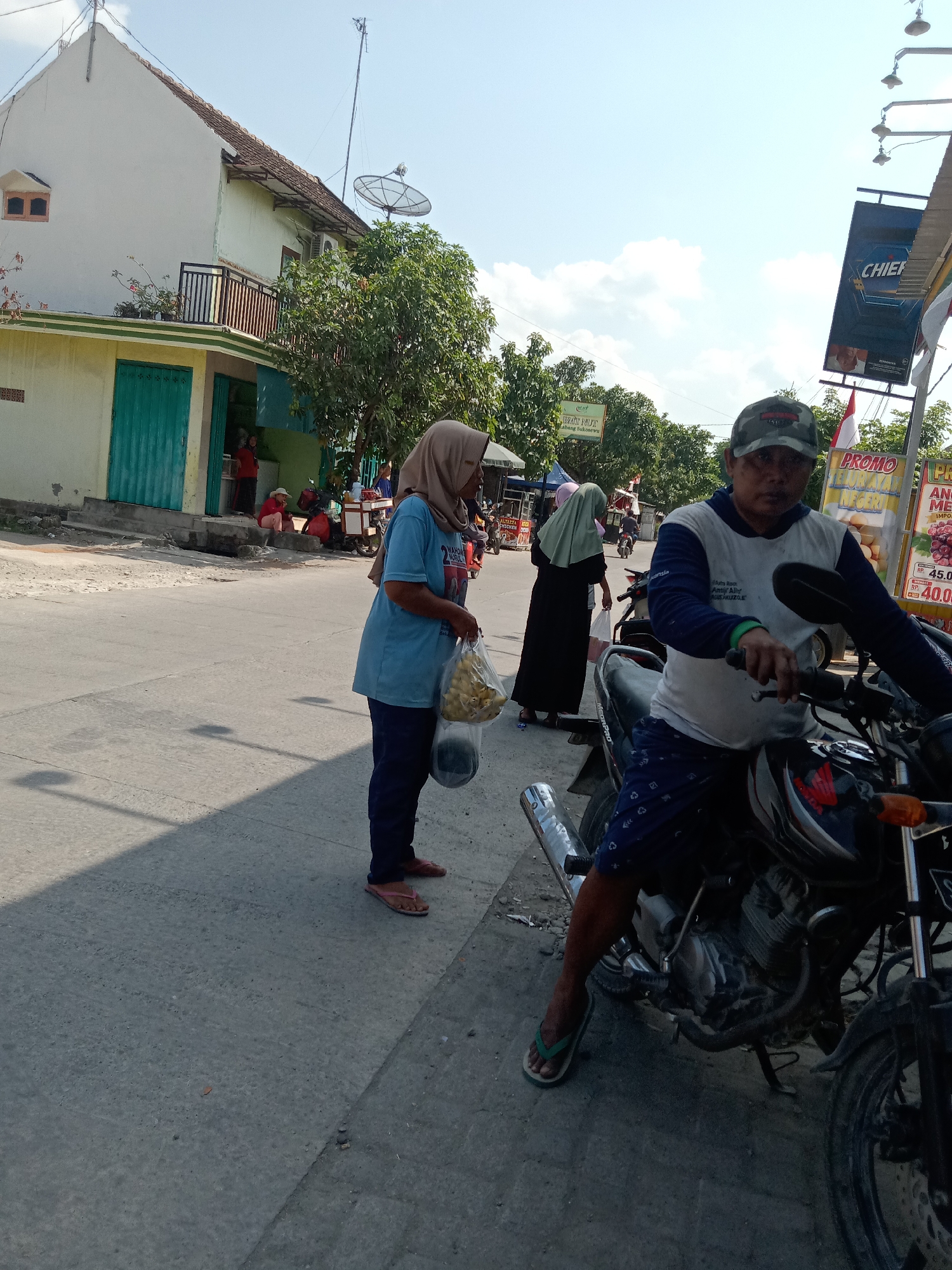 The image depicts a street scene in Klepek, Indonesia, under a clear blue sky with scattered clouds. A man wearing a camouflage baseball cap and a blue and white long-sleeved shirt is seated on a motorcycle in the foreground, facing the camera. He is wearing blue shorts with a white pattern. To his left, a woman in a light blue t-shirt and dark blue pants, wearing a beige headscarf, is standing and holding a plastic bag filled with yellow objects. She is also wearing flip-flops. Further in the background, two other women are visible. One is wearing a black abaya and a light green headscarf, and the other is partially obscured. Several other individuals are seated or standing near buildings and a cart in the middle ground. The buildings appear to be shops or residences, with one having a green door. A small tree is also visible. In the upper right of the image, there is a sign with "CHIEF" written on it, and a smaller sign with "PROMO" and some text in Indonesian. Another sign to the left displays "NYAMYI NUT." The ground is paved with asphalt. The overall scene suggests a bustling street environment.