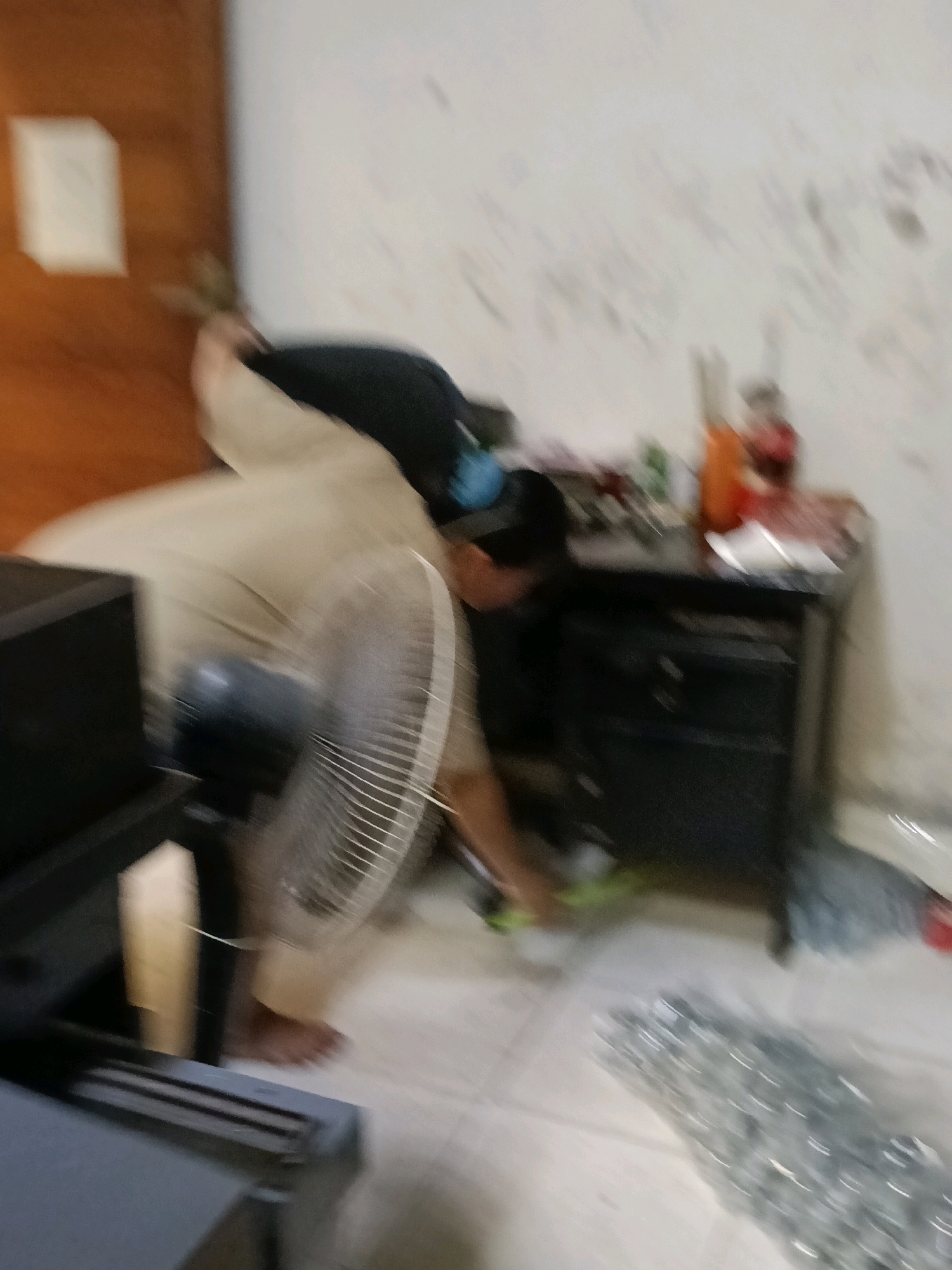 A person is bending over and appears to be cleaning or tidying an area. They are wearing a light-colored top and dark bottoms. A fan is positioned to their right, and a black cabinet with drawers is behind them. The room appears to be a home environment, possibly in the city of Belimbing, Indonesia. The walls are white and have some marks or writing on them. There are various items on top of the cabinet, including bottles. The floor is a light color.