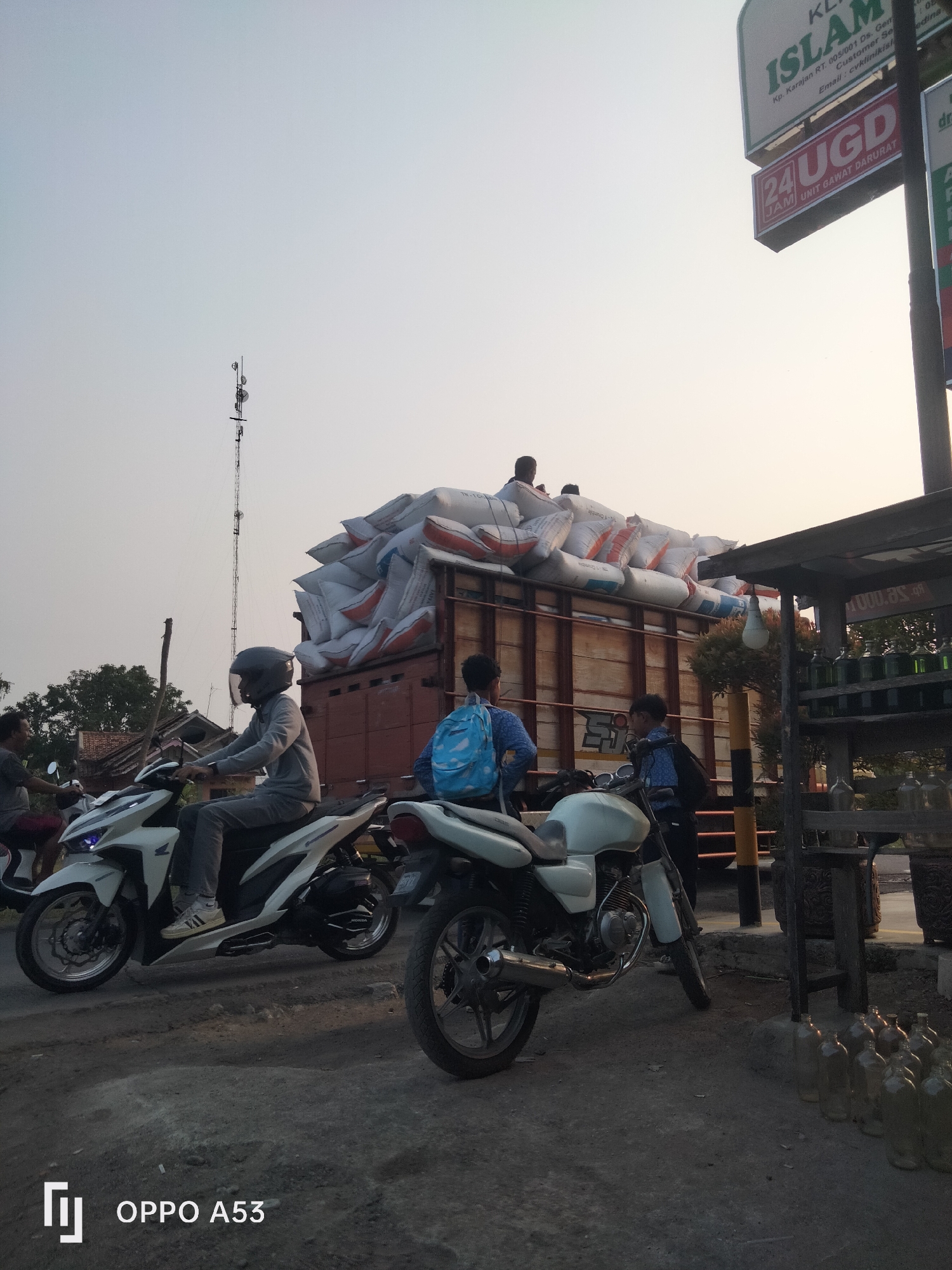 In Karawang, Indonesia, a street scene unfolds under a pale sky. A large truck is heavily laden with numerous white sacks, piled high and secured on its flatbed. Several people are visible, some standing on top of the sacks, while others are near the truck and its cargo. In the foreground, two motorcycles are parked. One is a white scooter-style motorcycle, with a rider wearing a helmet and grey long-sleeved shirt and pants. Another motorcycle, a white standard motorcycle, is positioned to the right. A person wearing a backpack is standing next to it. Further to the right, a wooden structure with shelves holds several glass bottles, some filled with a yellowish liquid, suggesting a roadside stall. Behind the truck, a tall communication tower stands against the sky. The road surface appears rough and worn. Signs are visible on a pole to the right, including one that reads "ISLAM" and another with "24 UGD JAM".