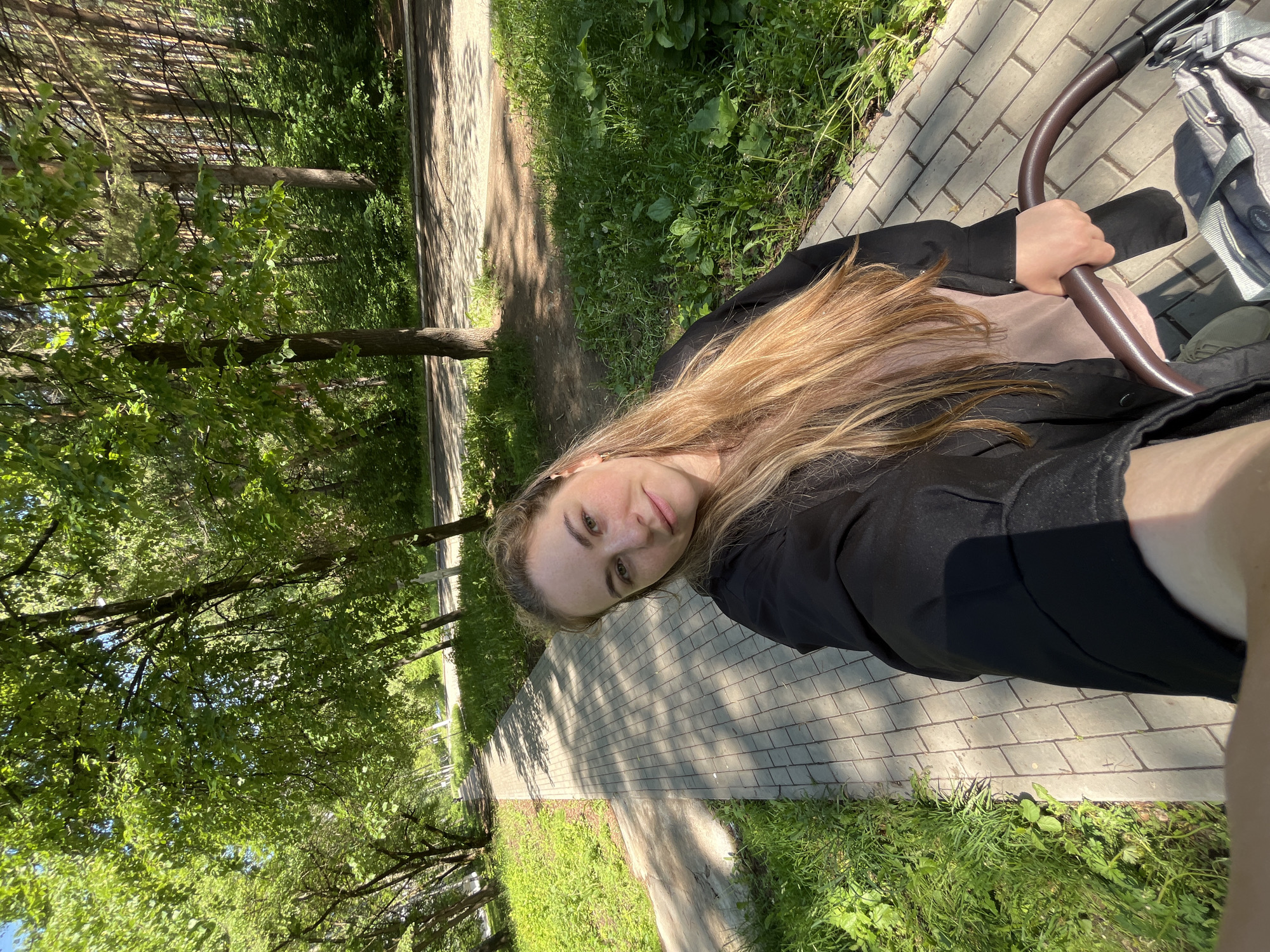 A young woman is captured in a selfie-style photograph, rotated 90 degrees counter-clockwise from its intended orientation. She has long, light brown hair, fair skin with freckles, and a calm expression with a slight smile. She wears a black top or jacket over what appears to be a light pink layer. Her right hand grips the dark brown, curved handle of a stroller or baby carriage, a gray portion of which is visible next to her.
The setting is an outdoor paved pathway in what appears to be a park or tree-lined area in Chaikovsky, Russia. The pathway is constructed from light-colored rectangular pavers and is flanked by vibrant green grass and numerous trees. Some trees exhibit lush green deciduous leaves, while others appear to be coniferous, with slender trunks reaching upwards. Bright sunlight illuminates the scene, casting distinct shadows of trees and foliage onto the pathway, indicating a clear, sunny day, likely in the morning or early afternoon. The overall impression is of a leisurely walk in a pleasant, green environment. There is no visible text in the image.