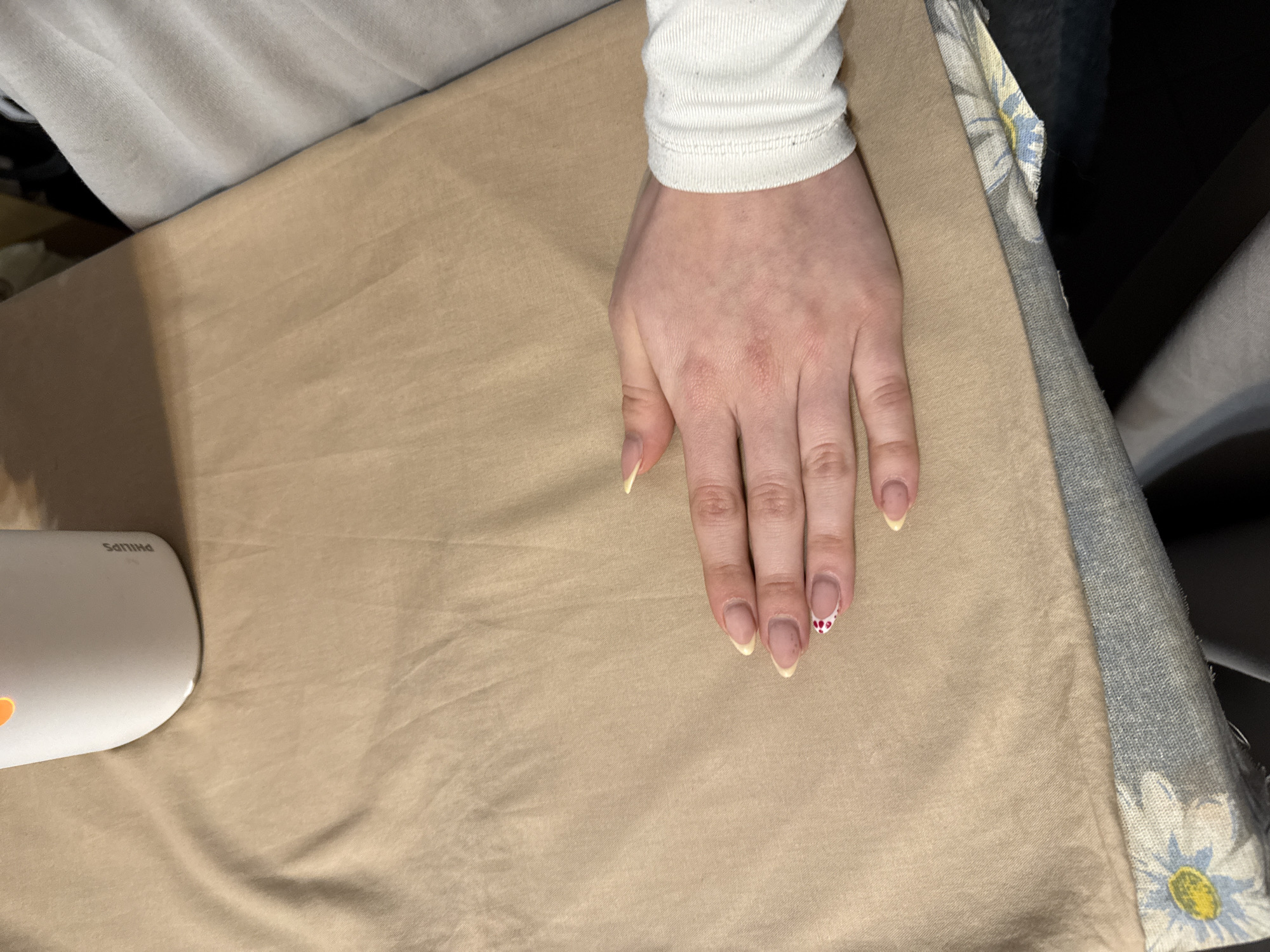 A right hand rests on a light brown surface, possibly a cloth or ironing board, in a brightly lit indoor environment. The hand belongs to a person wearing a white long-sleeved shirt. The fingernails are long and appear to have a light yellow-toned manicure, with one fingernail featuring a design of small red dots. A white Philips appliance is located on the left side of the frame. The right edge of the frame reveals a patterned fabric with a daisy design in grey, white, and blue. The location of the image within Sofia, Bulgaria cannot be determined from the visual information provided.