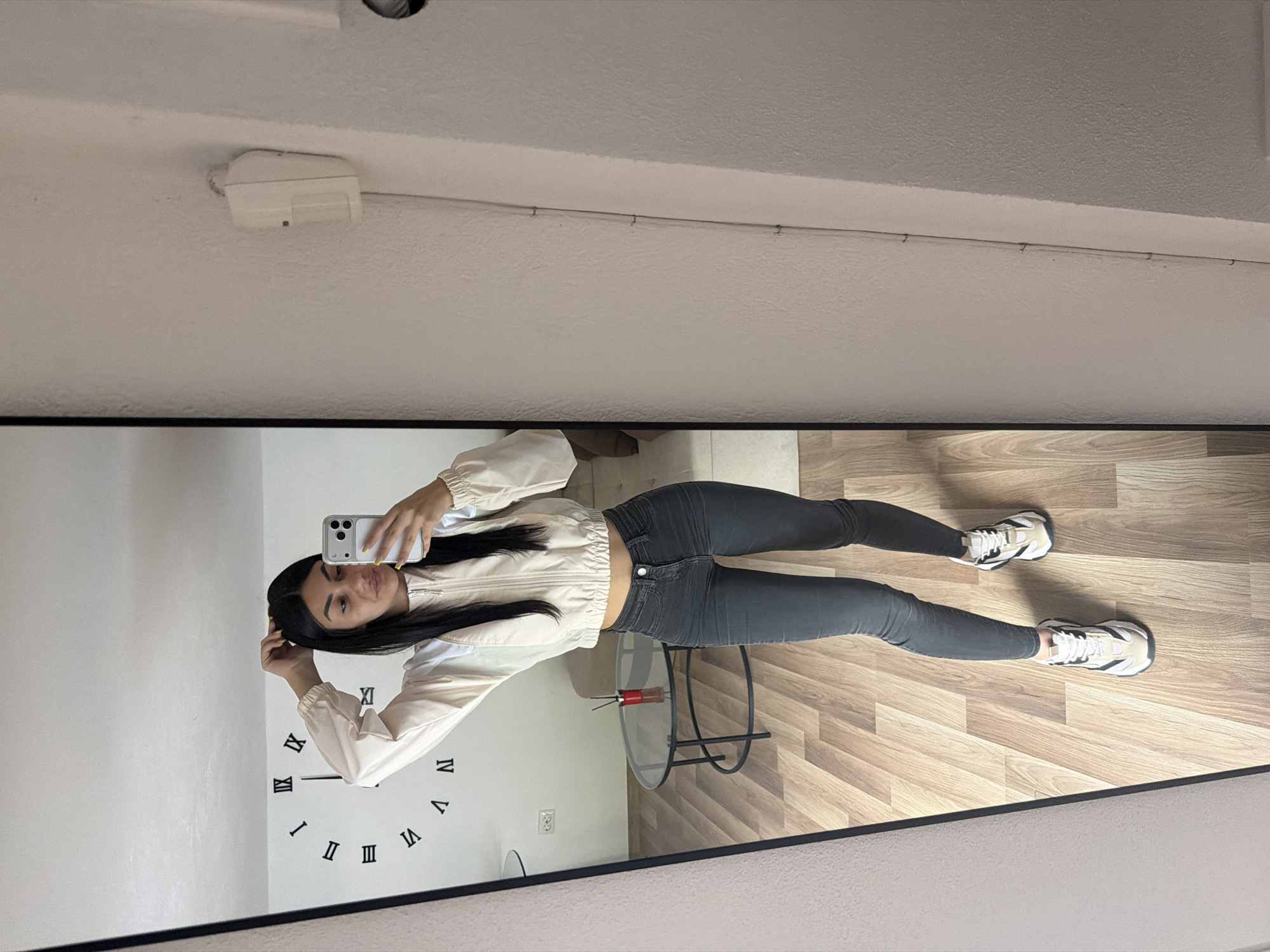 A young woman with long, dark hair takes a selfie in front of a large, rectangular mirror. She wears a light-colored jacket and dark jeans with white sneakers. A clock is mounted on the wall behind her to her left, with roman numerals marking the hours. A small round table is visible to her right. The mirror reflects a hardwood floor and a white ceiling with a white electrical fixture. There is no specific evidence within the image to pinpoint the location in Gabrovo, Bulgaria.