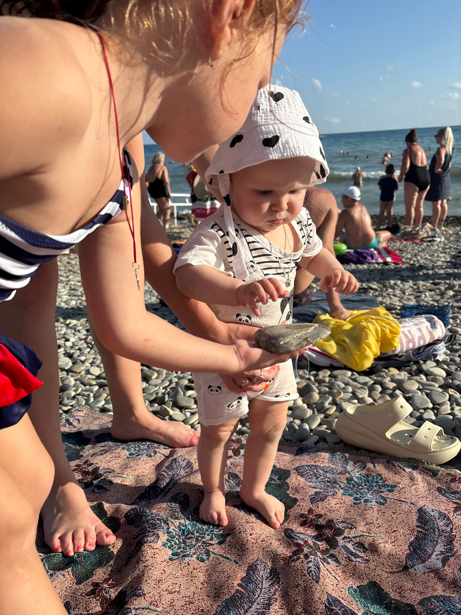 In a sun-drenched, outdoor scene, a young child wearing a white bucket hat adorned with black hearts and a romper with panda motifs is held by a larger child. The larger child, partially visible from the chest down, wears a navy and white striped bikini. They are both focused on a grey, oblong stone held in the larger child's hand, presented to the baby. The setting is a pebble beach, with the sea visible in the background under a clear blue sky with scattered clouds. Several other people are visible in the background, some wading in the water and others sitting on the shore. A colorful blanket with a floral and feather pattern is spread on the pebbles, with a pair of light-colored clogs placed on it. The overall impression is of a relaxed family outing on a warm day. The time of day appears to be daytime, likely mid-morning or afternoon, given the bright sunlight casting shadows and illuminating the scene. There is no visible text in the image. The location is Arkhipo-Osipovka, Russia.