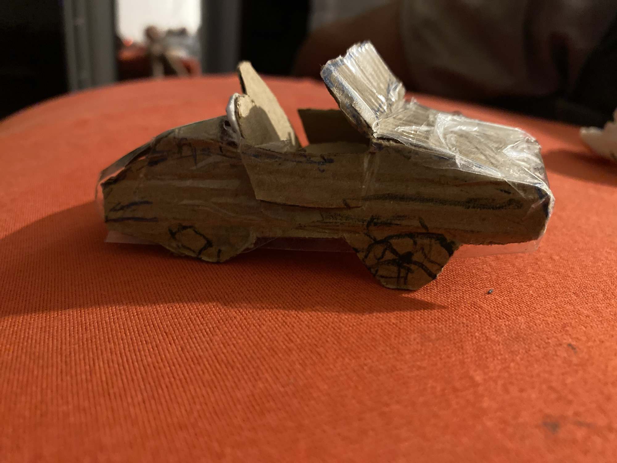 A handmade cardboard car toy rests on an orange textured surface. The car is brown and appears to be covered in clear tape. It has been decorated with black marker lines, suggesting it is meant to resemble a vehicle with wheels, doors, and a windshield. The surface it sits on has a subtle ribbed texture and is brightly lit, casting a shadow from the car. The background is blurred and dark, with hints of other objects or people, but they are not in focus.
