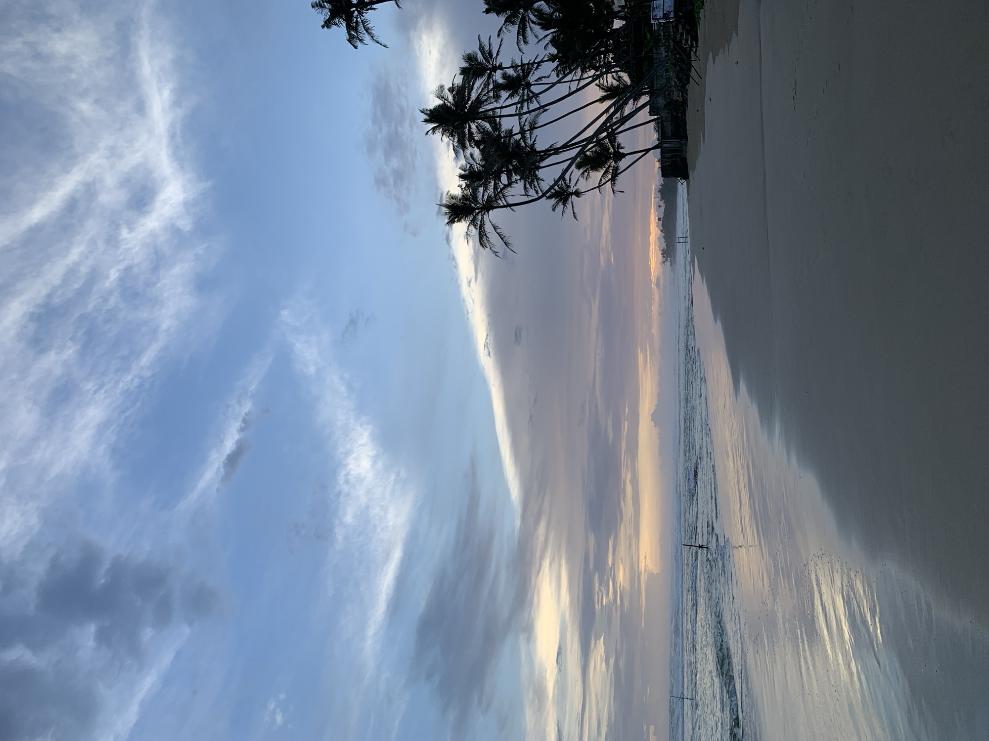 The image captures a beach scene in Kathaluwa, Sri Lanka. Palm trees line the shore, silhouetted against a sky filled with clouds in shades of blue, gray, and hints of orange and yellow from the setting sun. The sand is wet, reflecting the sky above. A few indistinct figures are visible in the distance. The water laps gently onto the beach.