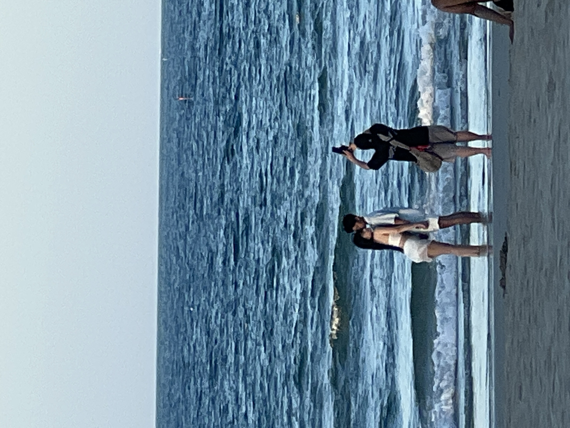 The media file shows a beach scene in Đà Nẵng, Vietnam. The image features two couples on a beach. One couple is standing in shallow water, with one person holding the other in a romantic pose. The other couple is further up the beach, with one person taking a photo of the other. The ocean is a prominent feature, with waves gently breaking on the shore. The sky appears clear and bright, suggesting it is daytime. The lighting indicates it is likely late afternoon or early evening, with the sun casting a soft glow on the scene. The overall mood is peaceful and idyllic, capturing a moment of leisure and connection by the sea. There is no visible text in the image.