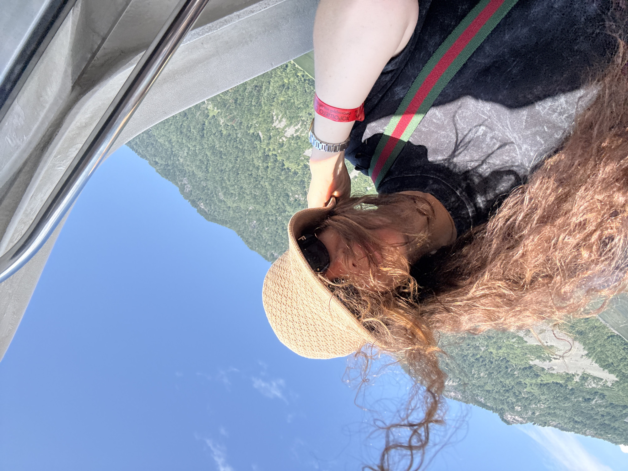 A selfie is taken by a person wearing a straw hat and sunglasses, with their hair blowing in the wind. The person is wearing a dark t-shirt with green and red stripes across the chest and a red wristband on their left arm. In the background, a lush green mountain range is visible against a clear blue sky. The person appears to be on a boat or other mode of transport, indicated by the metallic railing in the upper right corner of the frame. The location is the city of Kordon Baigazan, Russia.