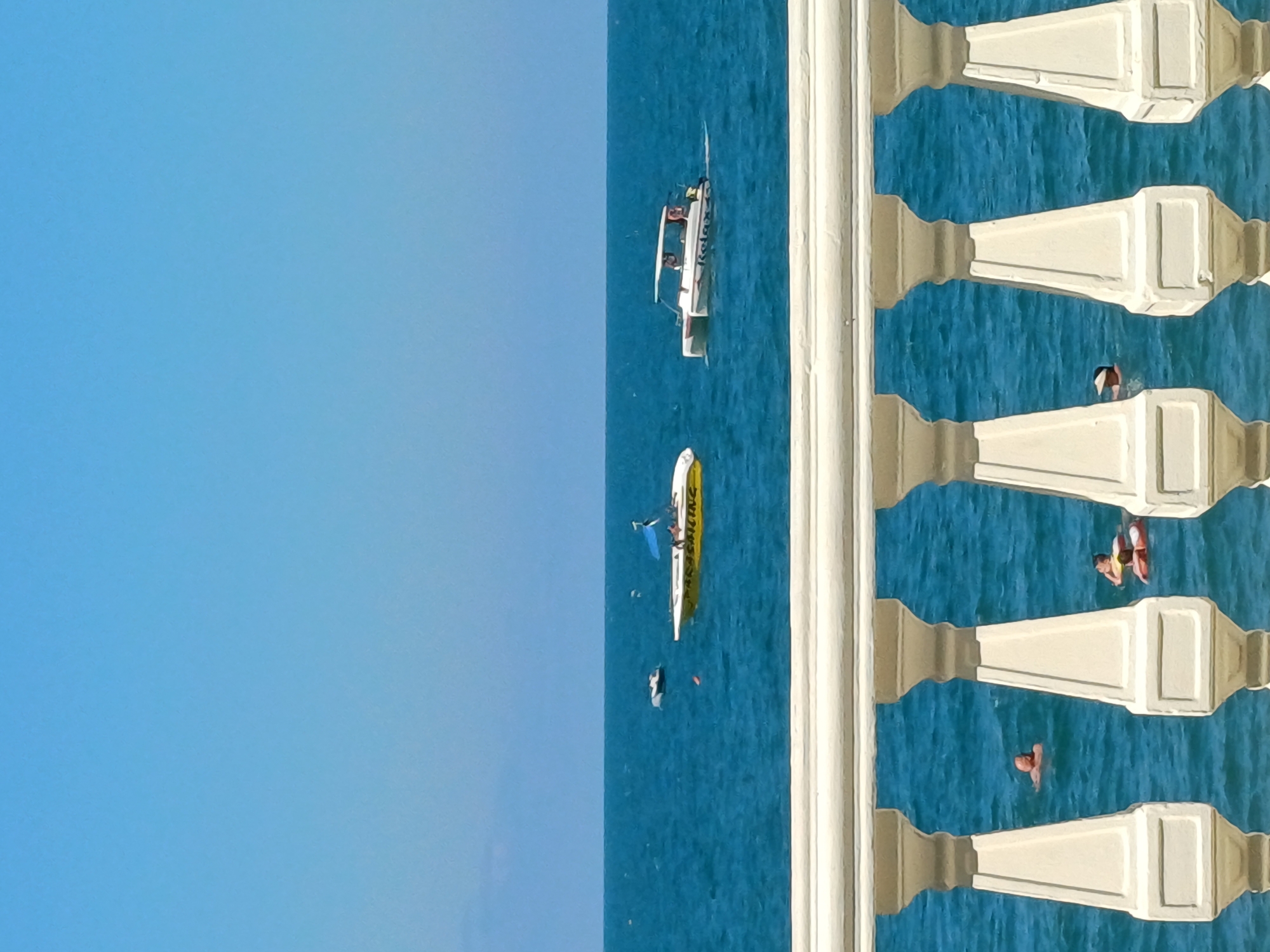 This media file depicts a scene from Sochi, Russia, overlooking the Black Sea. The image is oriented vertically, with the sky occupying the left half and the sea, along with a white railing, on the right.
In the sea, several objects and individuals are visible. There are two boats: a white speedboat with people on board, and a yellow and white parasailing boat. Several individuals are in the water, some appearing to be swimming or engaged in water activities. One person is on a flotation device.
The setting suggests a sunny day with clear blue skies and calm blue water. The white railing in the foreground, with its ornate balusters, provides a sense of being on a balcony or promenade.
Text is visible on the yellow and white boat, appearing to read "PARASAILING".
The overall impression is one of leisure and recreation, likely during daylight hours given the bright illumination.