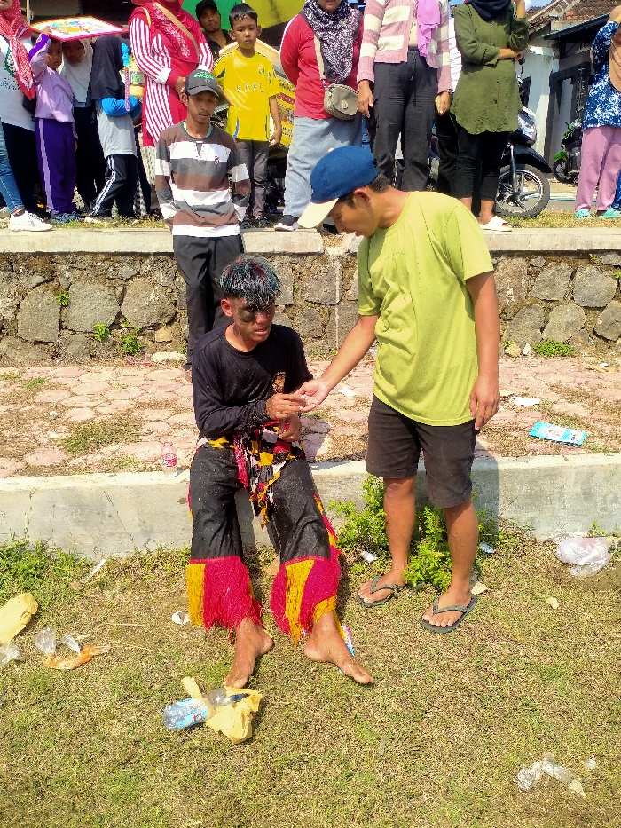 An outdoor scene in Celep, Indonesia, depicts two males in the foreground with a crowd observing from a higher elevation. In the immediate foreground, a male in his late teens or early twenties is seated on a concrete ledge. He wears a black long-sleeved shirt and black pants with red and yellow fringed fabric at the lower legs; the clothing appears wet. His face is smudged with a dark substance, and his hair exhibits multiple colors. He is barefoot, with his left hand extended towards the second male. This second male, appearing to be in his twenties or thirties, stands to the right, facing the seated individual. He wears a lime green t-shirt, black shorts, dark flip-flops, and a blue cap with a beige brim. His right hand is extended, interacting with the first male's left hand. The ground around them consists of sparse grass and dirt, littered with plastic bottles and wrappers. Behind the concrete ledge, a diverse crowd of approximately 10-15 individuals, including adults and children, stands on a paved surface. Many adults wear traditional head coverings. A black motorcycle is partially visible in the far background behind the crowd. The scene is brightly lit by daylight.