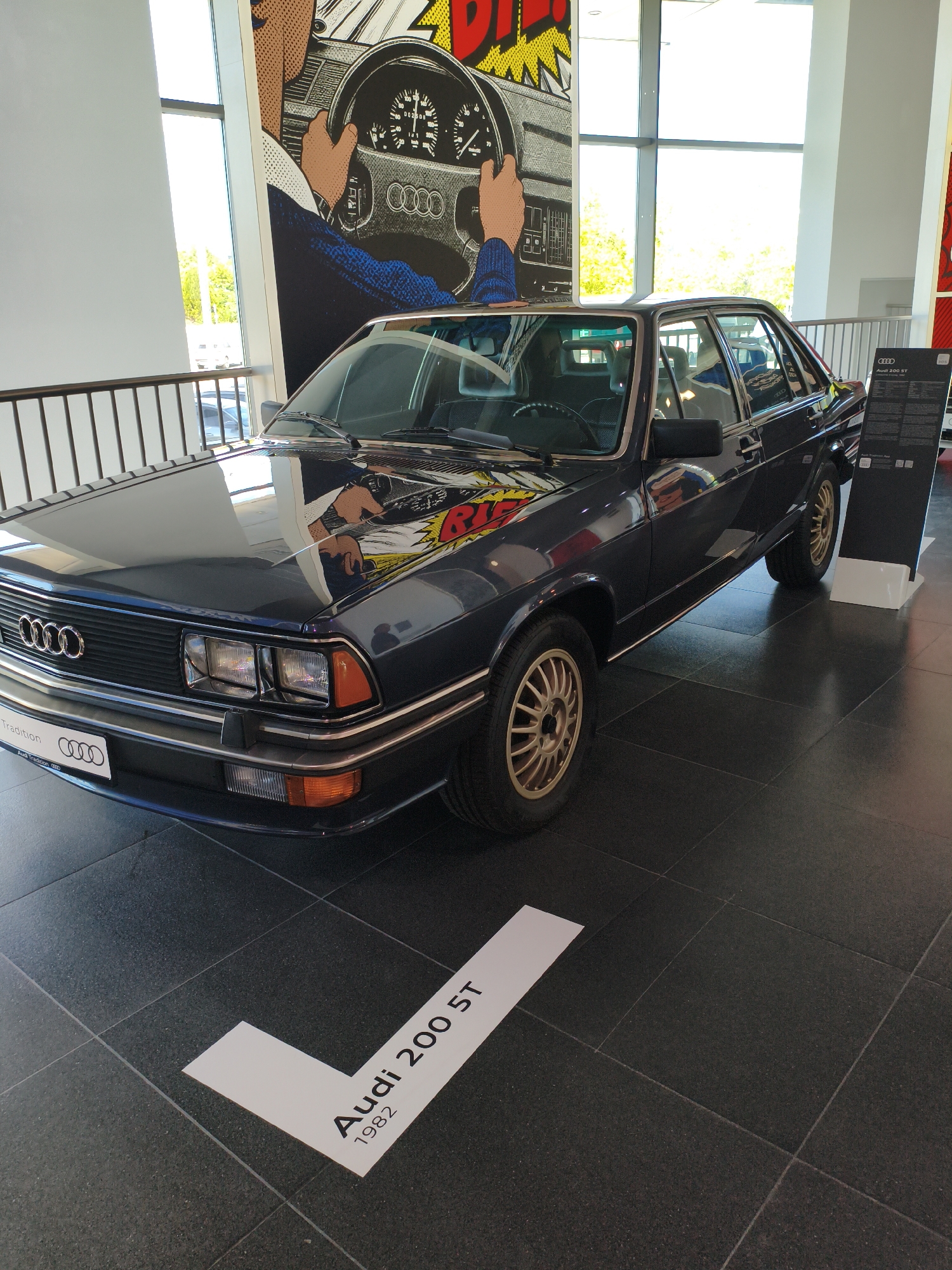 This is a photograph taken in the Audi Forum in Ingolstadt, Germany. The main subject is a dark blue 1982 Audi 200 5T sedan displayed on a dark, tiled floor. The car is positioned at an angle, facing towards the viewer's left, with its front end slightly closer to the camera.
The car's exterior features a prominent Audi grille with four silver rings, rectangular headlights, and amber turn signals. The hood of the car reflects a large, colorful pop-art style mural depicting a driver's hands on a steering wheel. The wheels are a distinctive gold color with a multi-spoke design.
On the floor in front of the car, a white directional graphic points towards the vehicle. This graphic has the text "Audi 200 5T" and "1982" printed in black. To the right of the car, a black display board with information about the car is visible, along with the Audi logo.
In the background, large windows allow natural light to enter the space, illuminating the scene. A metal railing runs along the left side of the frame. The overall environment appears to be a modern exhibition space, likely a showroom or museum dedicated to Audi automobiles. No people are visible in the photograph.