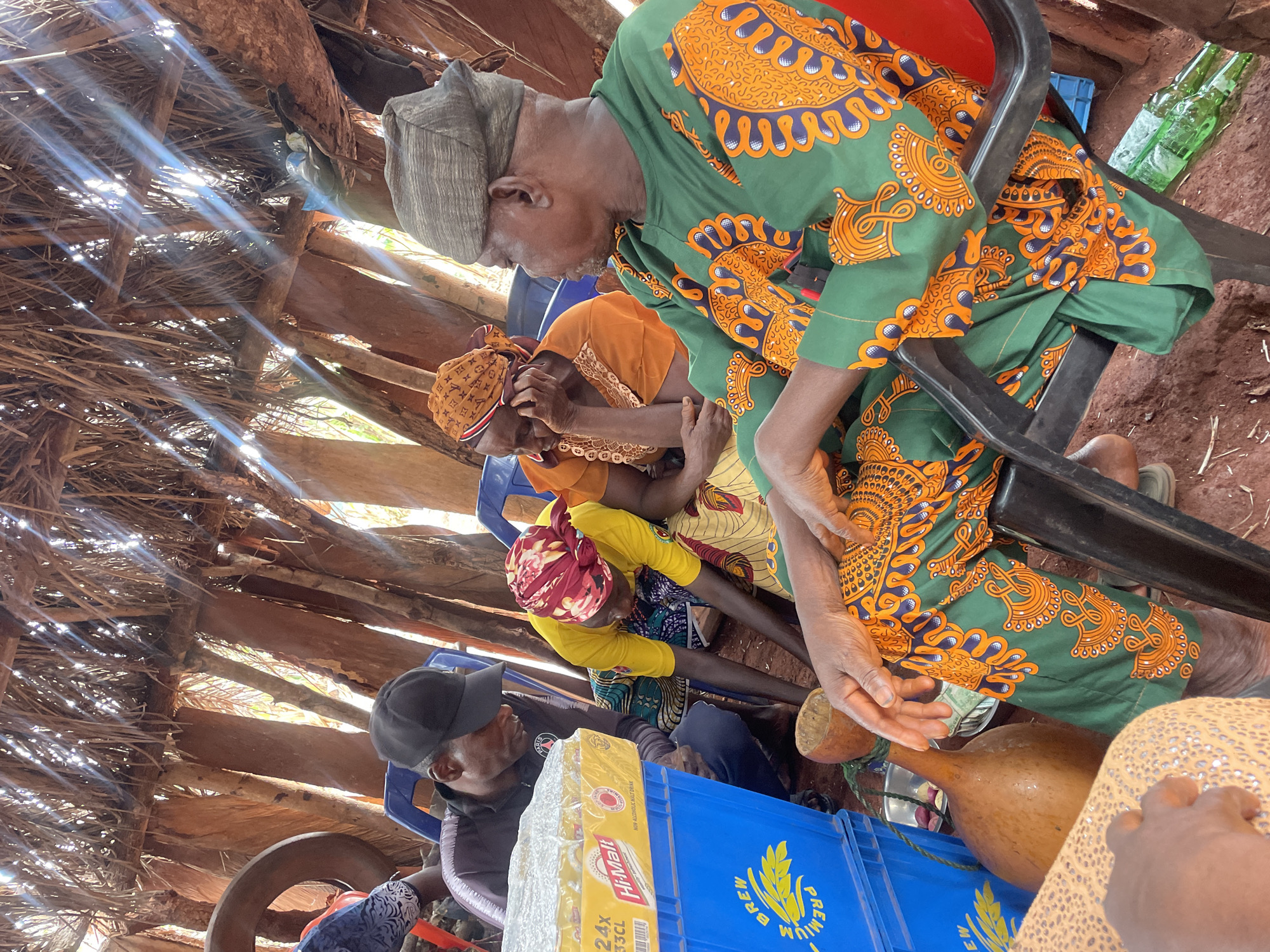 The scene depicts several people inside a structure with a thatched roof, presumably in Ogbe, Nigeria. Four adults are seated on plastic chairs. The first adult wears a green and orange patterned garment. The second adult wears an orange outfit. The third adult wears a yellow top. The fourth adult, seated to the far left, wears a black cap and jacket. A blue crate of "Hi-Malt" drinks rests on the floor in front of him. Between the individuals are several gourds of various sizes. The floor appears to be red soil. The ceiling is made of thatched roofing material with gaps letting in sunlight.
