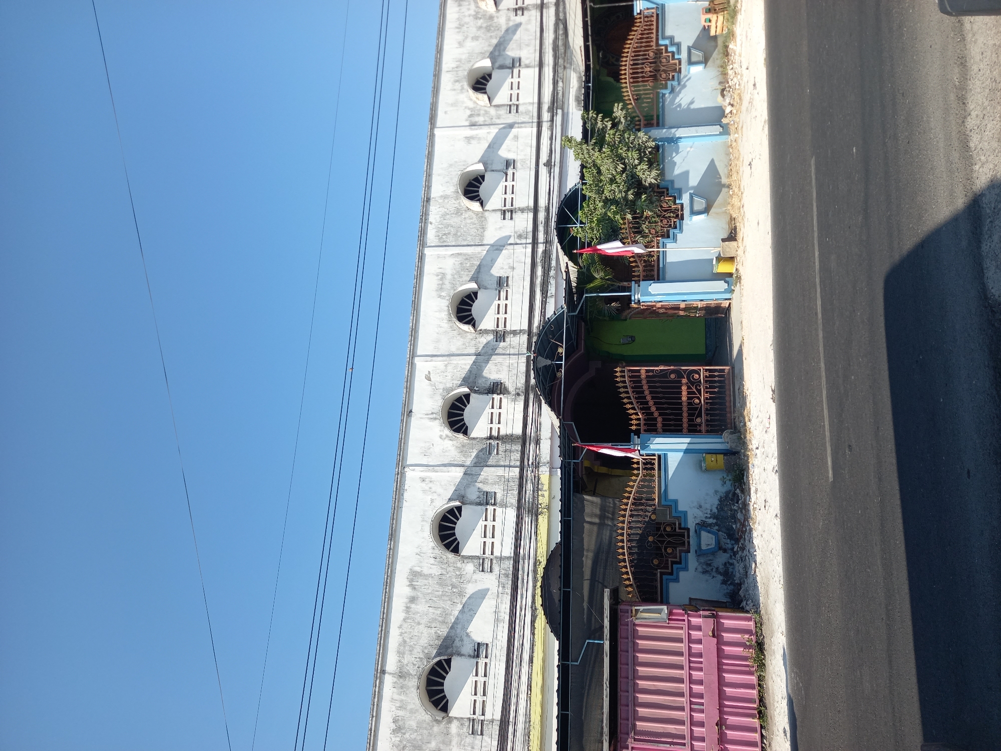 The image shows a clear blue sky above a building with a textured white facade. The building features a row of arched windows with segmented panes, casting shadows on the wall. Below the building, a metal fence with intricate scrollwork separates the property from the street. To the left, a pink corrugated metal structure stands. An Indonesian flag is planted near the gate, and a green bush grows in front of the fence. The street in front of the property is asphalt, with a faint white line marking the center. The scene appears to be an outdoor view on a sunny day.