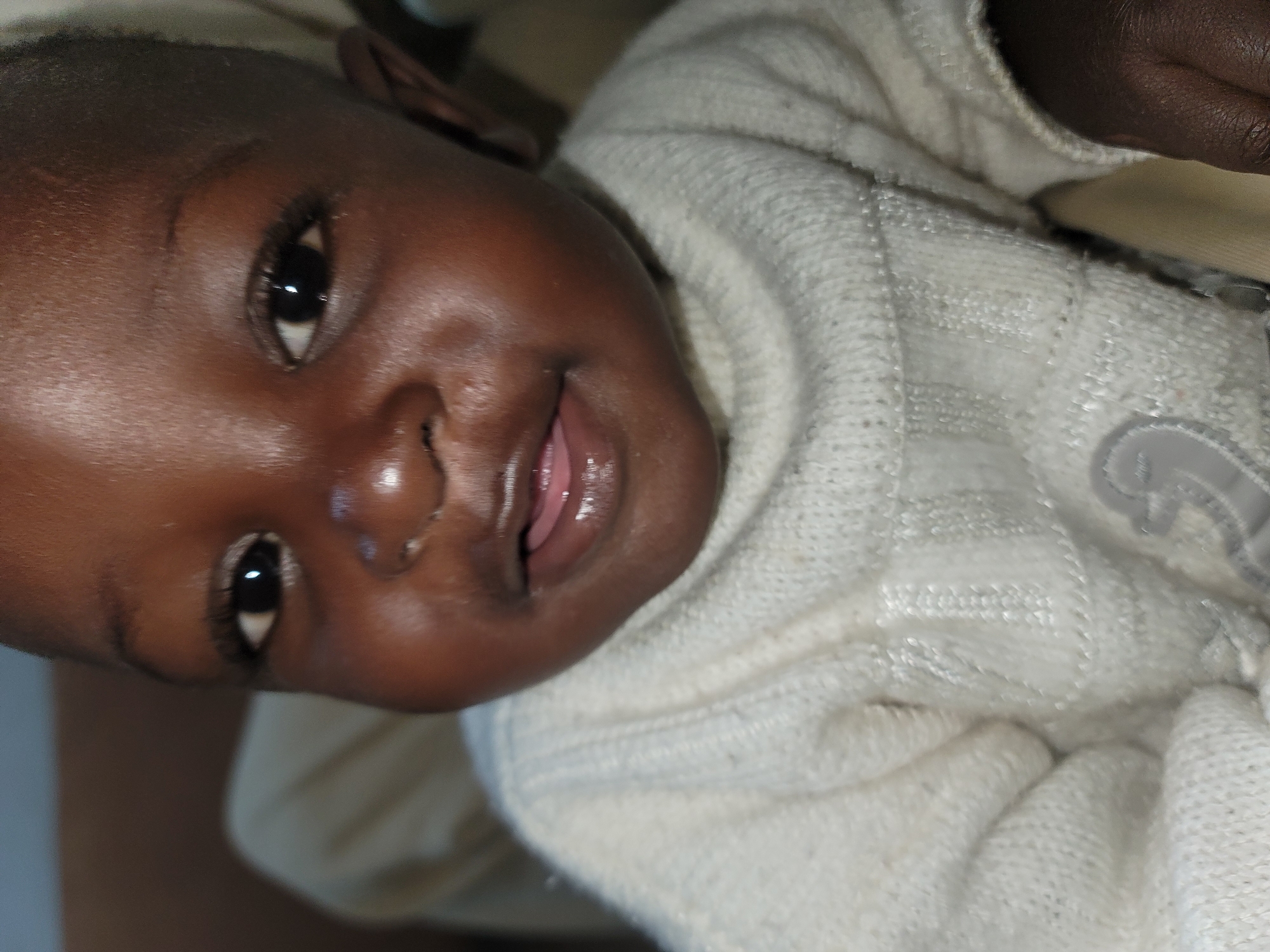 A close-up, eye-level, indoor, portrait-style photograph shows the face of a young Black child. The child is looking directly at the camera. The child has dark skin, dark hair, and dark eyes. The child is wearing a white, knitted turtleneck sweater. The background is blurred and appears to be an indoor setting with soft lighting. The image is in color and is sharp.