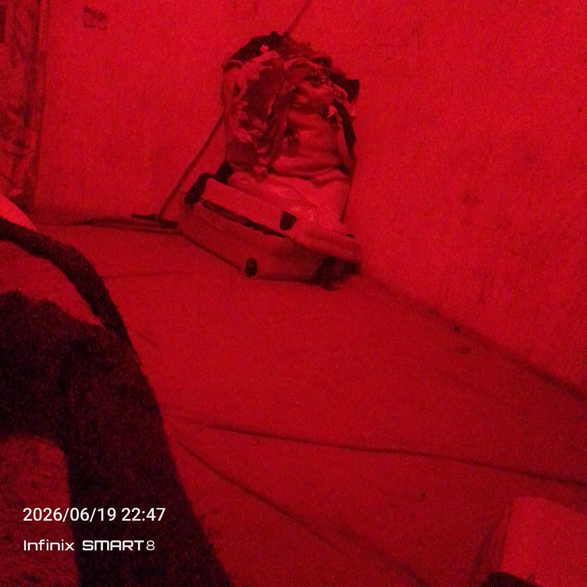 The image depicts an indoor scene with a heavy red lighting effect. In the background, a pile of clothes or fabric is placed on top of a white object, possibly a piece of furniture. The floor is a flat surface, possibly carpeted or tiled, with some visible lines or creases. A dark, textured object, likely a rug or blanket, occupies the left foreground. The bottom left corner of the image displays a timestamp and device information: "2026/06/19 22:47" and "Infinix SMART 8". The overall impression is one of a dimly lit, possibly domestic, environment. There are no people or discernible actions taking place. The location is noted as Unknown, Unknown.
