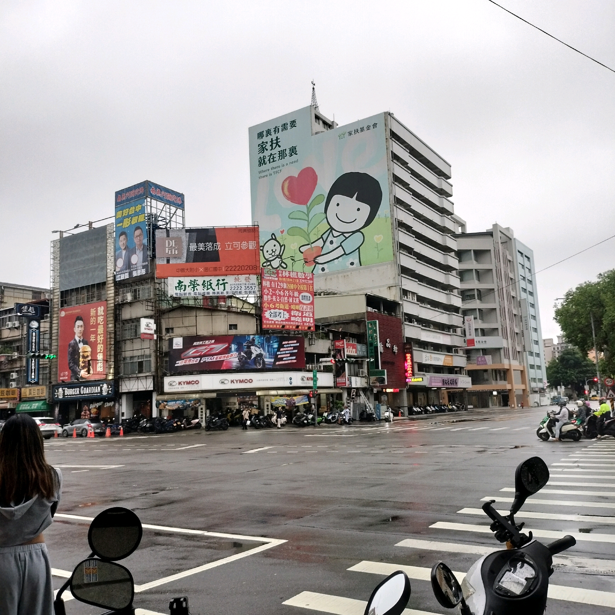 The image depicts a busy intersection in Taichung, Taiwan, likely on a cloudy or overcast day, as indicated by the muted lighting and wet road surfaces suggestive of recent rain. Prominently featured are several tall buildings adorned with large advertisements and billboards. One billboard showcases a cartoon child holding a heart-shaped flower, with text in Chinese and English reading "Where there is a need there is TFCC." Another billboard displays two men in suits, possibly politicians or businessmen. Below these, various storefronts are visible, including "BURGER GUARDIAN" and "KYMCO" dealerships, with motorcycles parked in front. Several individuals are present; one woman in a grey tracksuit stands in the foreground, facing away from the camera, looking towards the intersection. Other people are seen on the sidewalks and riding motorcycles. The intersection itself has a pedestrian crosswalk, and traffic lights are visible. The overall scene is characteristic of a commercial and urban area.