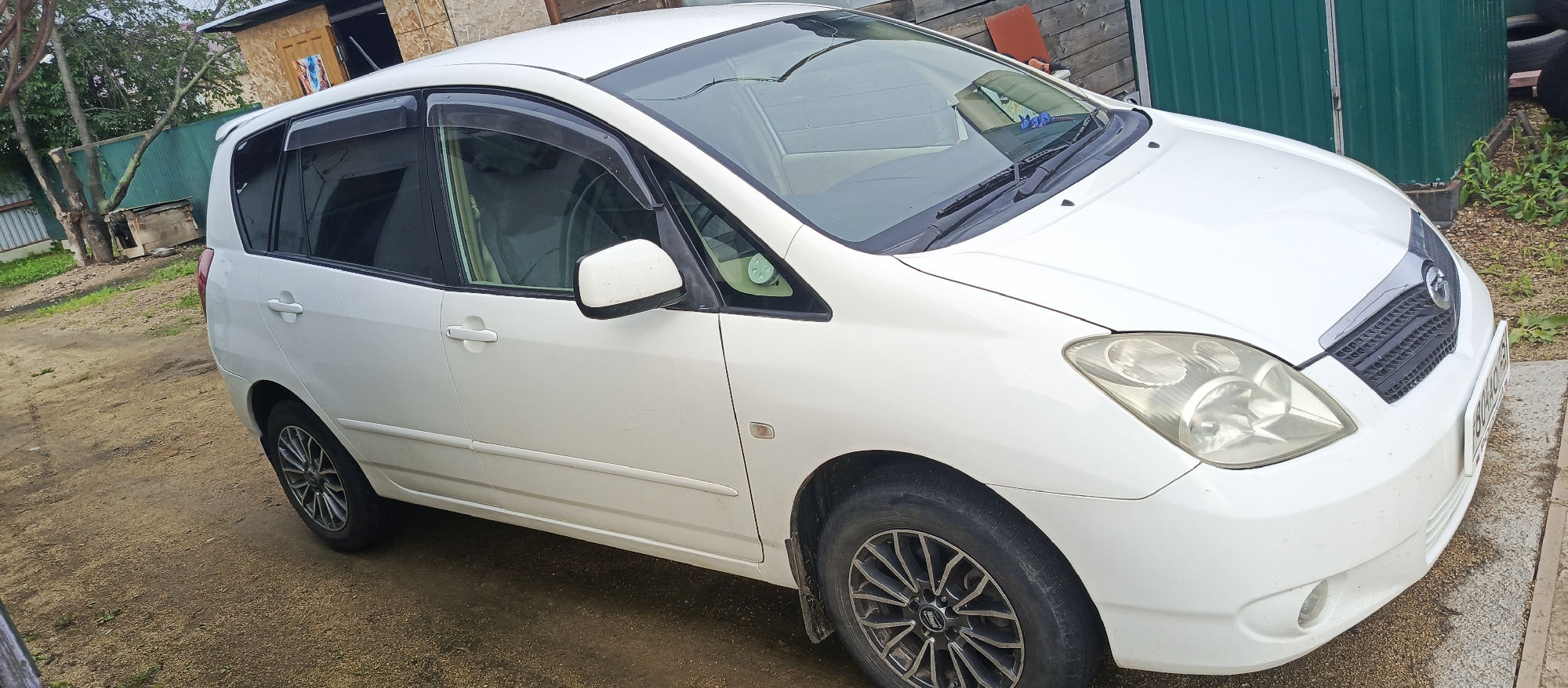 A white Toyota Ipsum is parked on a dirt lot next to a weathered wooden building. The car has tinted windows and aftermarket rims. The building appears to be an outdoor shed or storage unit. In the background, there is a green metal fence and some overgrown vegetation. The scene is dimly lit, suggesting it might be overcast or late in the day. There are no people or animals visible in the image. The car's license plate is visible, displaying Russian characters and numbers. The location is the city of Pogranichny, Russia.