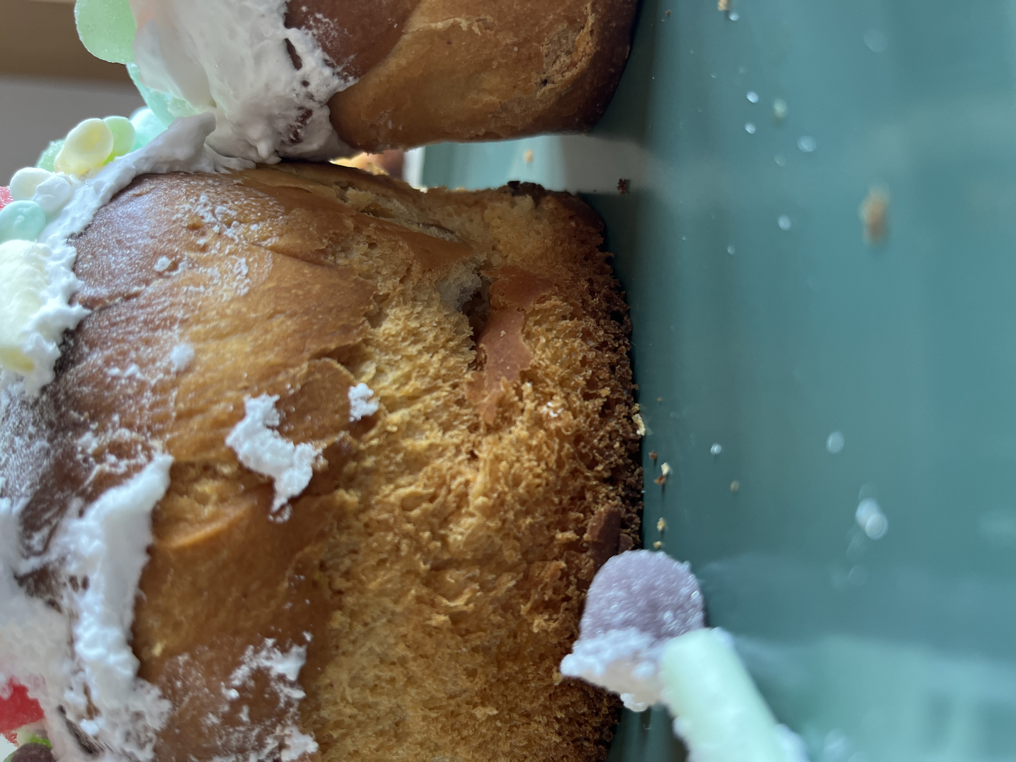 A close-up, slightly tilted, indoor shot shows a decorated Kulich bread. The bread is covered in white icing and pastel-colored candies. The bread is sitting on a pale green plate. Debris is visible on the plate.