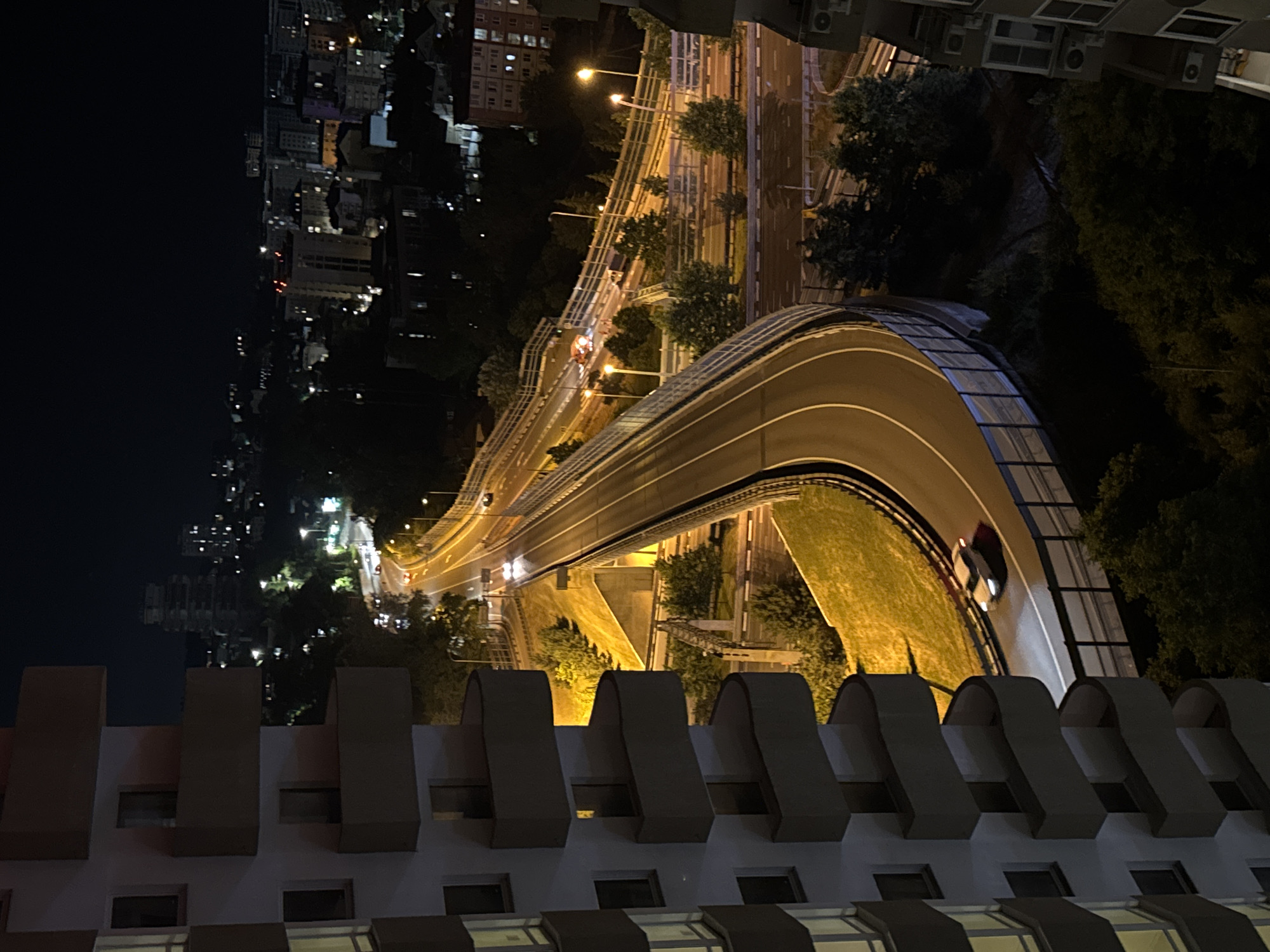 An elevated view at night captures a highway interchange in Sochi, Russia, with a multi-story building in the foreground on the left. The highway curves through the scene, illuminated by streetlights and the headlights of vehicles. One car is visible on the highway, appearing white. Further up the highway, a cluster of emergency vehicles, including a white van and possibly an ambulance or fire truck with flashing lights, are present, suggesting an incident. In the background, a cityscape with numerous illuminated buildings and distant lights stretches across the horizon under a dark night sky. The building in the foreground is partially lit, revealing balconies and windows. The environment is characterized by artificial lighting and the dark expanse of the night.