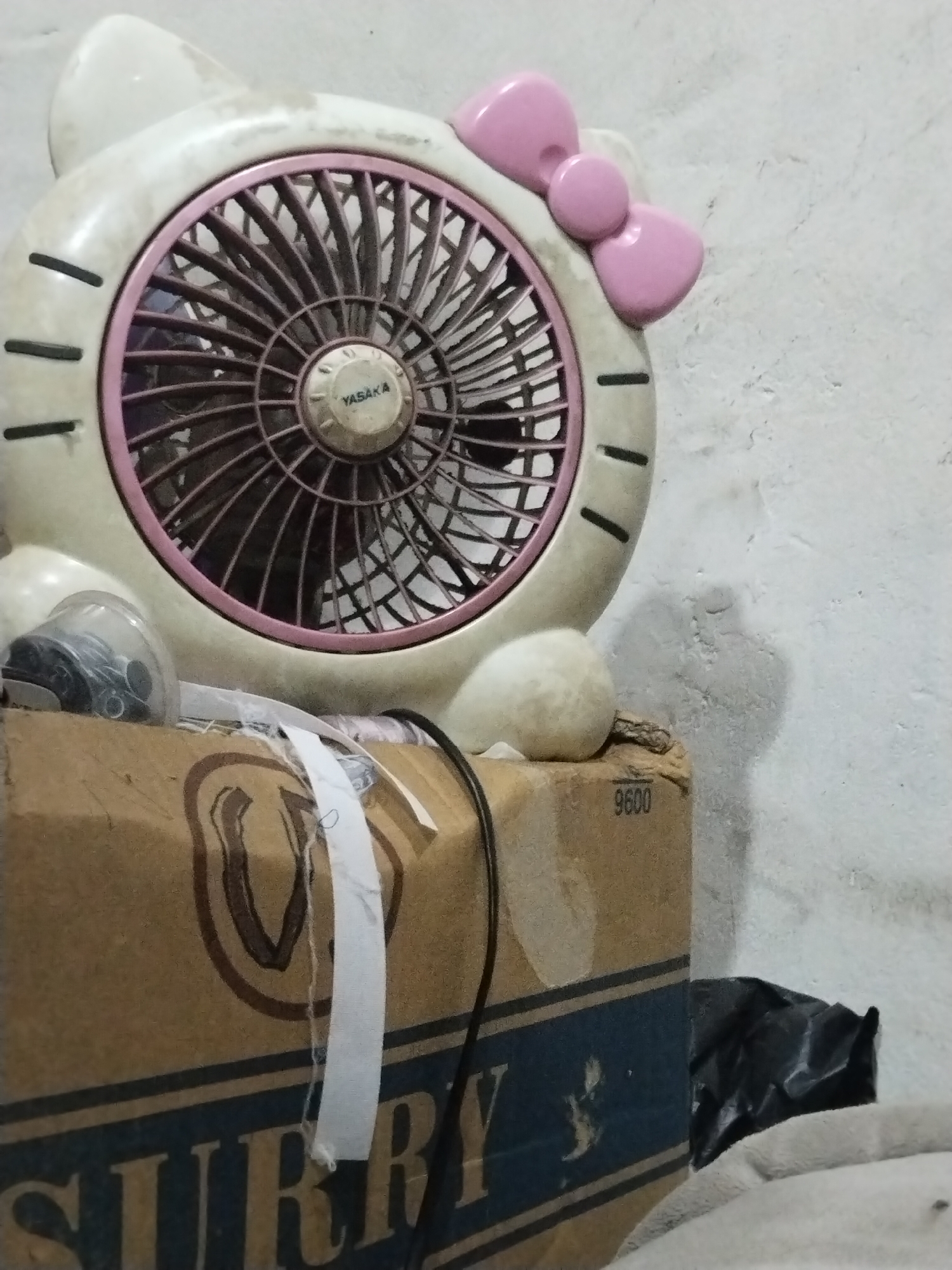 An indoor, eye-level, medium shot captures a Hello Kitty-themed fan positioned on top of a cardboard box. The fan, a cream-colored plastic casing with pink accents, features a circular grill and a pink bow on its head. The fan is angled towards the left, with its blades visible through the grill. To the left of the fan, a clear plastic object with coiled wires is partially visible.
The fan rests on a brown cardboard box, which is the primary foreground element. The box has a dark blue stripe along its bottom edge, with white lettering that is partially obscured but appears to read "SIRRY" in a block font. There are also brown markings and a white tape with some blue string running across the top of the box. A black plastic bag and a light-colored blanket are visible behind the box and to the right of the fan, respectively.
The background consists of a textured, light-colored wall, likely concrete or plaster, with some unevenness and minor marks. The lighting appears to be ambient and somewhat dim, casting a faint shadow of the fan to the right on the wall. The overall impression is one of a storage area or a domestic setting, possibly in a modest urban environment. There are no people or animals present in the image. The scene is static and appears to be undisturbed.