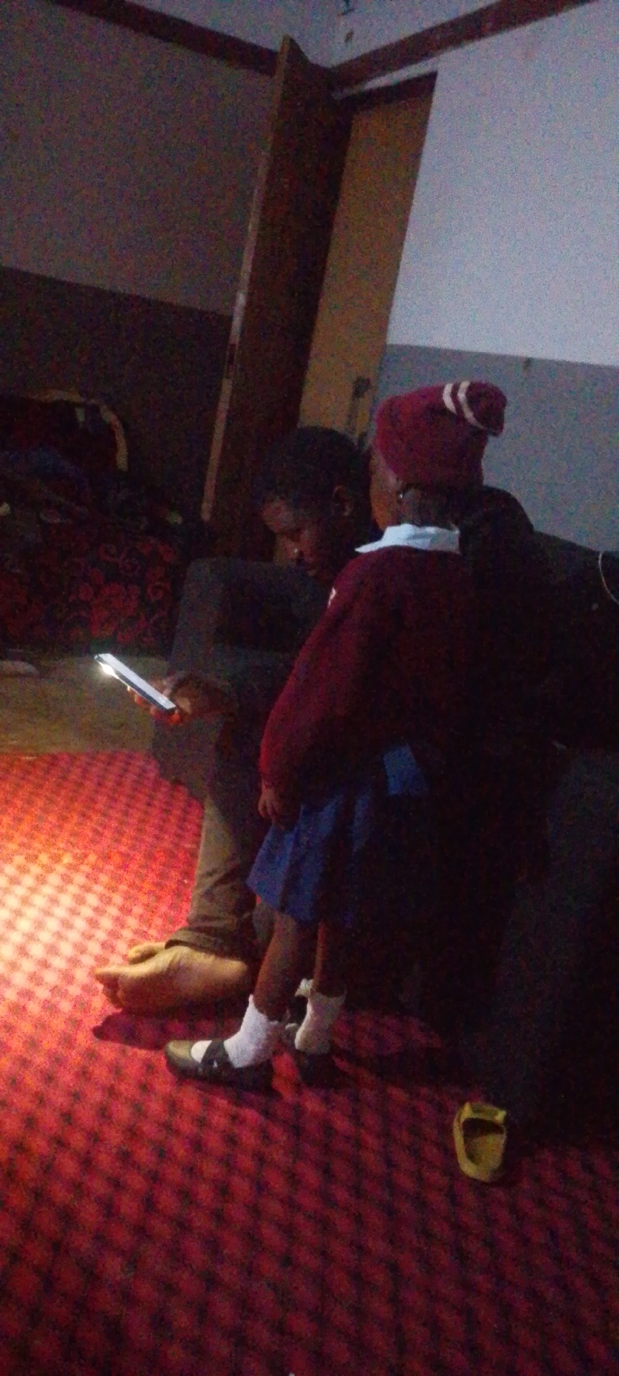 Two individuals, one adult and one child, are seated on a red carpeted floor. The adult is on the left, holding a lit smartphone and looking down at it. They are wearing dark pants and are barefoot. The child is on the right, wearing a maroon sweater with a white collar, a blue skirt, white socks, and black Mary Jane shoes. The child is also wearing a maroon hat with white stripes. The background appears to be a dimly lit indoor space with furniture and a wooden structure. There is a yellow object resembling a slipper on the floor near the child's feet.