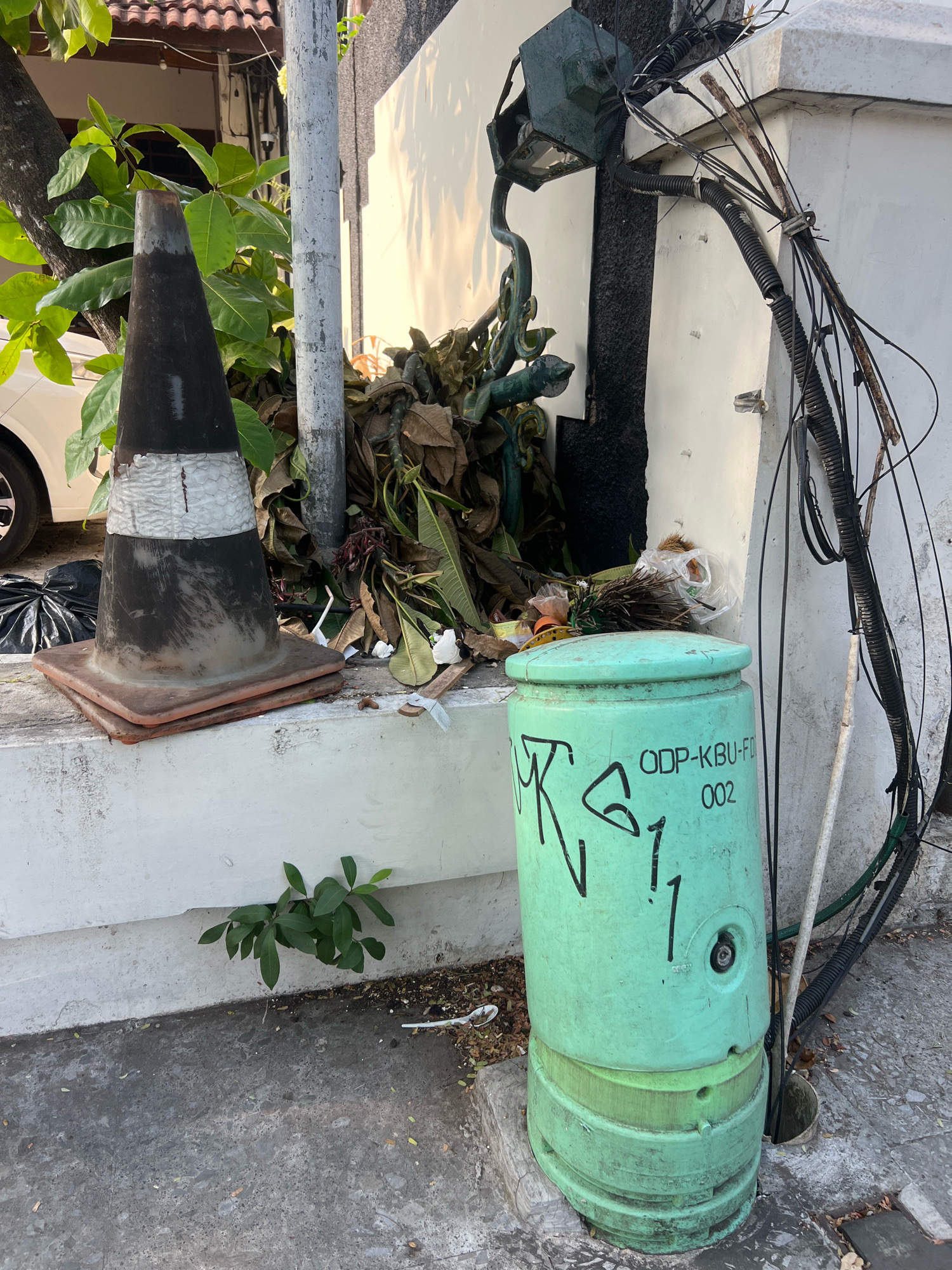 The image depicts an outdoor scene in Yogyakarta, Indonesia, characterized by urban detritus and infrastructure. In the foreground, a large, weathered orange traffic cone with black and white markings rests on a concrete ledge. To the right of the cone, a green cylindrical utility box, possibly for telecommunications or electrical equipment, is situated on the ground. It bears markings that appear to be graffiti or identification numbers, including "ODP-KBU-FD 002." The box is surrounded by loose debris, including dried leaves and plastic waste. Behind the utility box, a jumble of electrical cables and conduits are visible, attached to a white wall. A metal pole, possibly a streetlight or utility pole, stands next to the cone. A pile of dried leaves and branches is heaped against the wall behind the cone and pole. In the upper left quadrant, a portion of a white vehicle is visible, suggesting a street or parking area nearby. Lush green foliage from a tree is visible in the upper left. A small green plant is growing out of a crack in the concrete wall in the lower left portion of the frame. The overall impression is one of a neglected urban space with accumulated refuse and visible utility infrastructure.