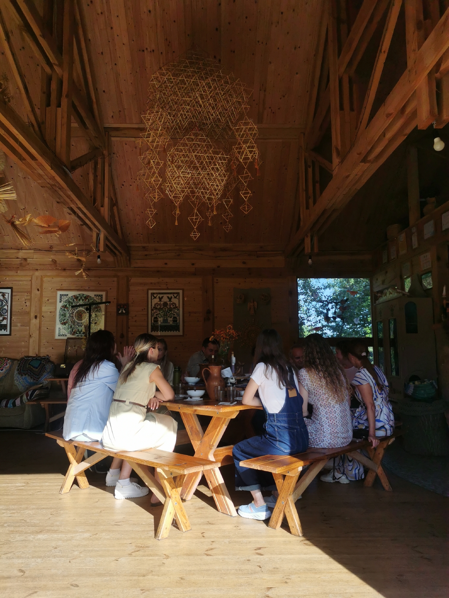 The image depicts an indoor gathering within a rustic wooden structure. Six individuals, viewed from behind, are seated around a wooden table. The room features exposed wooden beams and paneling, with a large, geometric hanging decoration suspended from the ceiling. On the walls, decorative artwork and other hangings are visible. The table is set with plates, bowls, and a pitcher. A projector screen displays an image of green foliage and possibly birds. The overall ambiance suggests a casual, communal event in a rural or artisanal setting. Based on the provided location, this scene is situated in Шчарчоўскі сельскі Савет, Belarus.