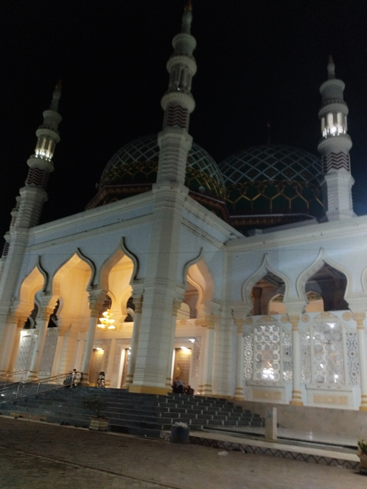The image displays a large mosque at night in Kotapantonlabu, Indonesia. The structure is predominantly white or light-toned, illuminated by internal and external artificial lighting. Two large, dark green domes, featuring a diamond grid pattern and gold-colored decorative elements at their bases, cap the main building. At least three minarets are visible; two flank the main structure (one on the far left, one on the far right), and one is centrally positioned between the domes. These minarets are cylindrical, white, and adorned with red and white zig-zag patterned bands; their upper sections emit light. The mosque's facade presents multiple pointed arches, through which a warm, yellow light is visible from the interior. Pillars support these arched sections. On the lower right side of the building, intricate white latticework panels are integrated into the wall design. A wide, multi-tiered staircase, dark grey in color and equipped with metal handrails, leads up to the mosque's entrance. Two indistinct human figures are discernible on the staircase. The foreground consists of a dark, unpaved surface, with a small potted plant situated near the base of the stairs. The sky above is completely dark, indicative of a nocturnal setting.