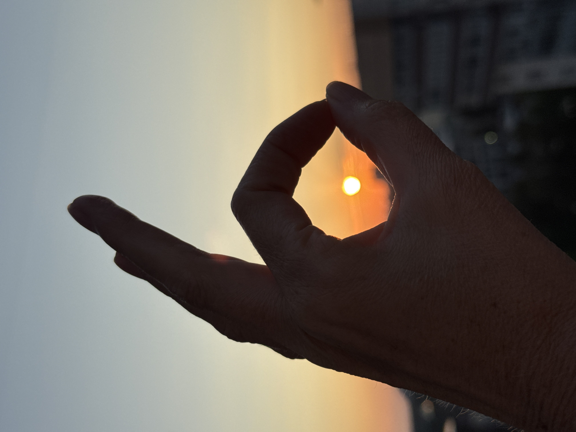 A silhouette of a hand forms an "OK" gesture against a twilight sky. The sun, a small, bright circle, is captured within the hand's loop, appearing as if it's being held. The background shows a blurred cityscape with the soft glow of the setting sun casting an orange and yellow hue. The sky transitions from a pale blue at the top to warmer tones near the horizon. The hand's skin texture is subtly visible in the silhouette. The image captures a moment where the sun is framed by the hand, creating an artistic composition. The exact location in the city of Unknown, Russia, is not identifiable from the visual information.