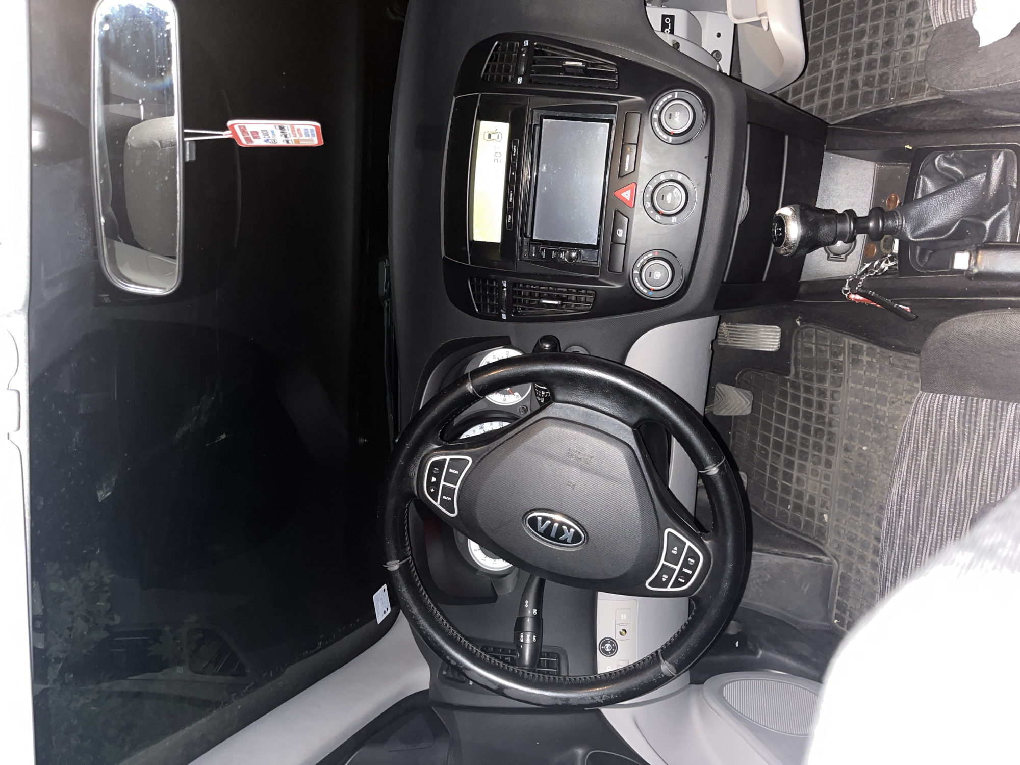 The image shows the interior of a Kia car. The steering wheel, dashboard with a display screen, and gear stick are visible. A price tag hangs from the rearview mirror. The car's interior is black and grey. The floor mats have a square pattern. The image provides no information about the location of the car in Marulevo, Bulgaria, nor does it depict any people or actions.