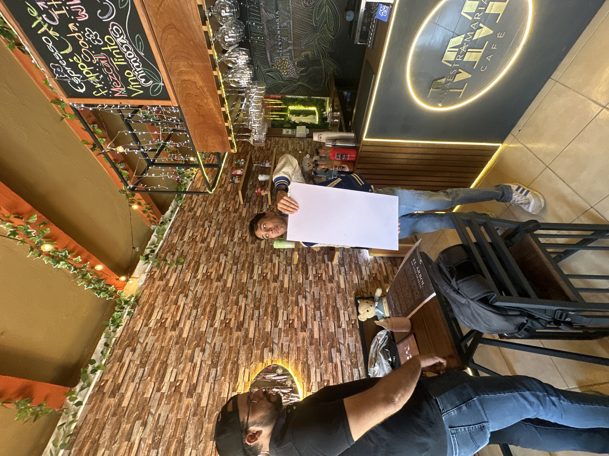 Here is a detailed description of the image:
**Scene:**
The image shows the interior of a café named "MAESTRA MARIA CAFÉ".
**Location:**
The café is located in the city of San Andrés Cholula, Mexico.
**People:**
There are two people visible in the image. One person is holding a blank white rectangular sheet. Another person is seated at a table.
**Subjects:**
The subjects include people and objects found within the café.
**Objects:**
Objects include:
- A blank white sheet
- A menu with the text "EL ARBOL DE LOS DESEOS"
- A backpack
- Chairs and tables
- Glassware
- A blackboard with a menu including items like "Espresso", "Frap Soda", "Mezcal", "Vino Tinto", and "mosas"
- Wall decorations, including a neon logo, plants, and a brick wall design
**Actions:**
One person is holding a blank white sheet, while the other is seated at a table.
**Interactions:**
There does not appear to be any direct interaction between the two people shown.
**Environment:**
The café's interior has a combination of brick and brown walls. The lighting is a mix of natural and artificial. There are bar and table seating areas.