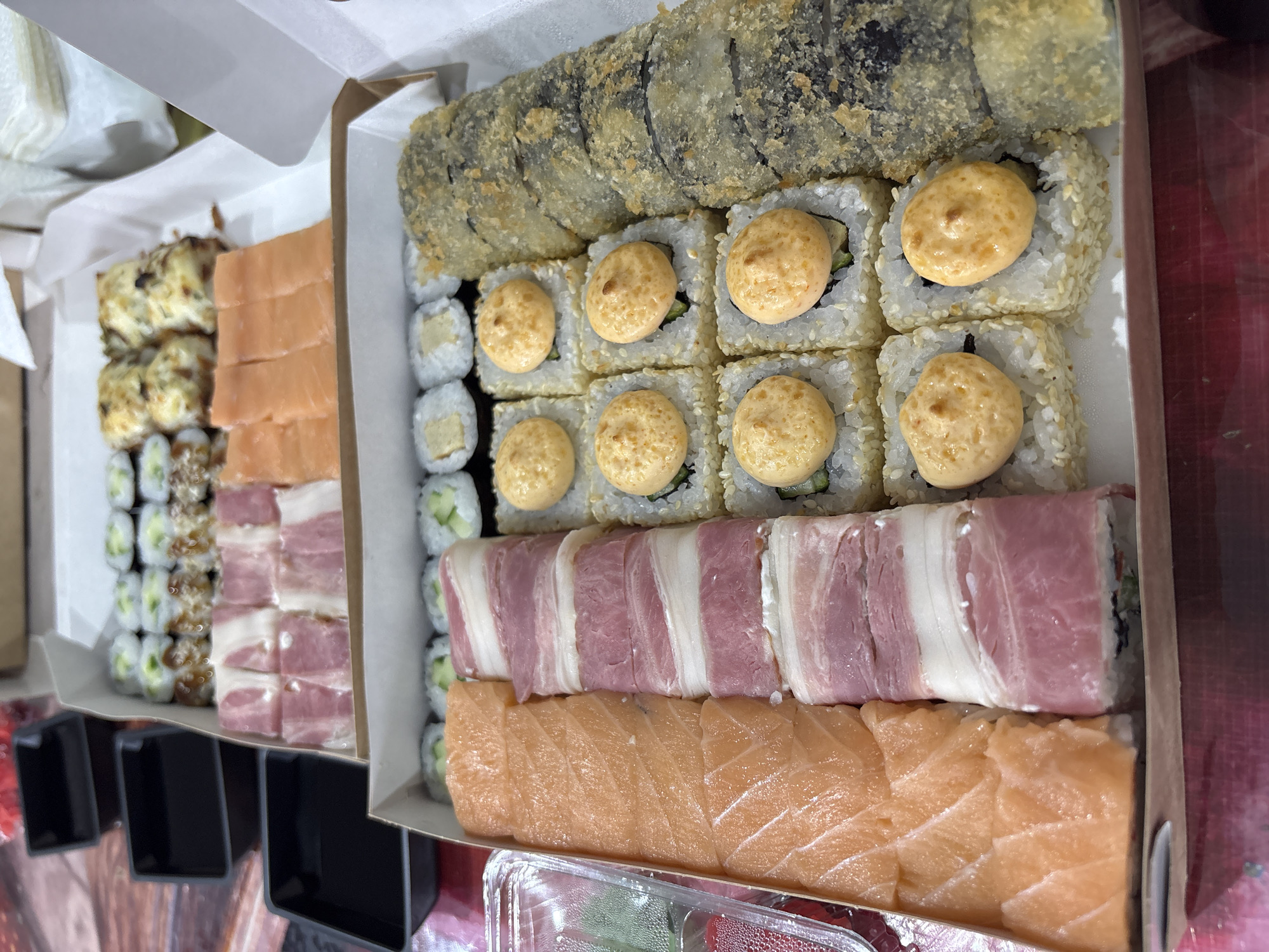 The image shows two cardboard boxes filled with sushi. The box on the left contains rolls with cucumber, rolls topped with fish and cream, and rolls with bacon. The box on the right contains a large fried roll, rolls topped with a creamy sauce, rolls with bacon, and rolls topped with fish. There is no location information in the image.