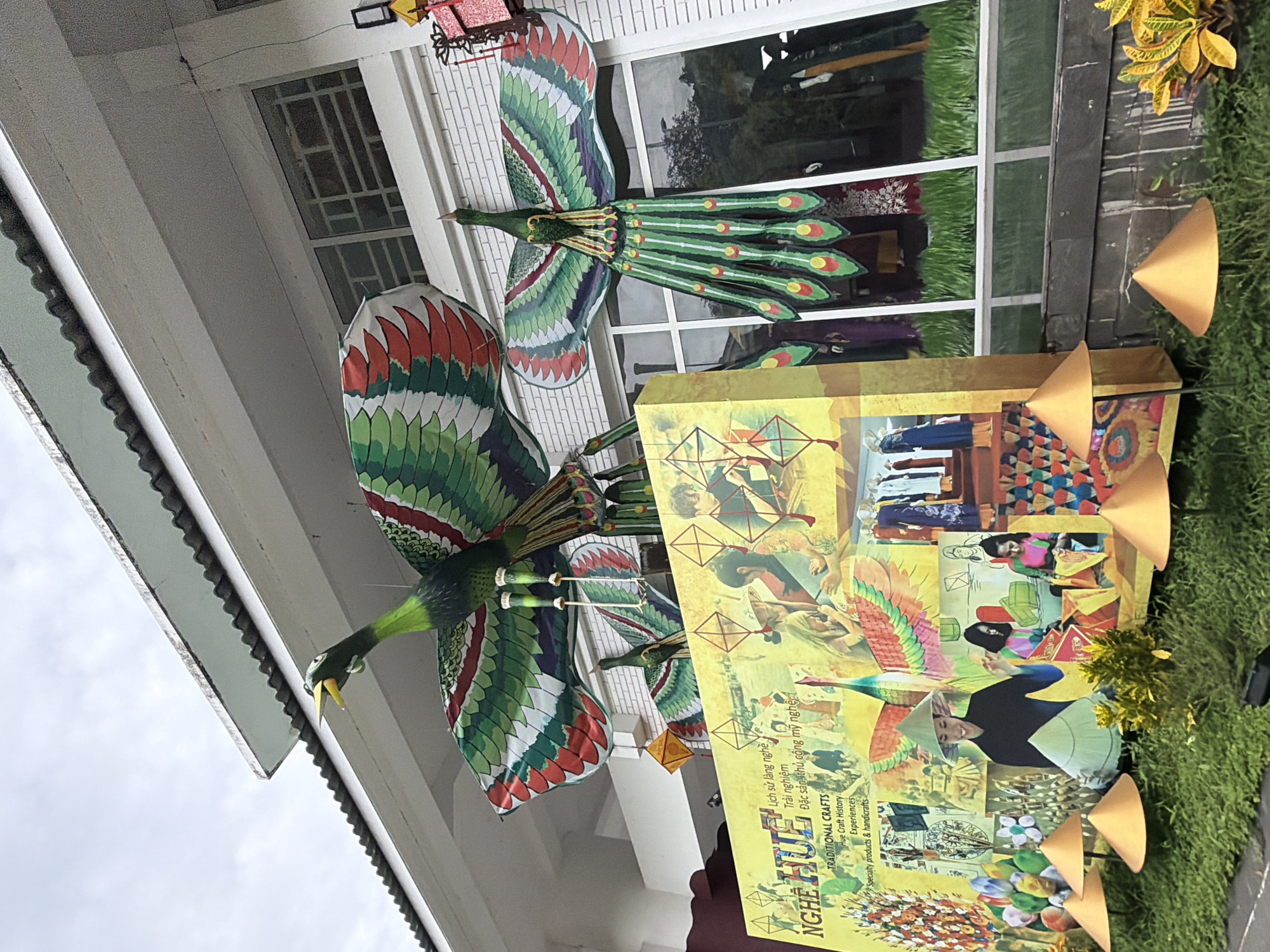 This image captures an outdoor display in Thuận Hóa Ward, Vietnam, featuring large, decorative kites shaped like birds, specifically peacocks. The kites are vibrant green, red, and white, with spread wings and elaborate tail feathers. They are suspended in front of a building with large windows, on which reflections of the surrounding environment are visible.
At the base of the display is a large, yellow rectangular structure adorned with images and text. The text includes "NGHỆ HUẾ" and "TRADITIONAL CRAFTS," suggesting the display promotes traditional crafts from the Hue region of Vietnam. The images on the structure depict scenes of people engaged in crafting, wearing traditional conical hats, and displaying colorful fabrics.
In the foreground, a lush green lawn is dotted with several golden, conical decorative elements. The overall scene suggests a cultural exhibition or a festive decoration, likely related to a local festival or a promotion of Vietnamese heritage and handicrafts. The sky above is overcast.