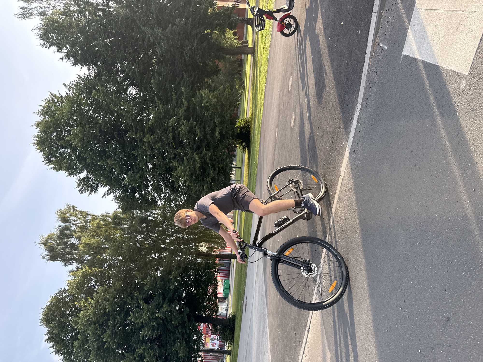 A young Caucasian male with blonde hair and sunglasses rides a black bicycle on an asphalt path. He is wearing a dark grey t-shirt and dark grey shorts. The bicycle has black handlebars, a black frame, and black tires with orange reflectors on the spokes. Behind the cyclist, a red electric scooter with a black basket is parked. The scene is set in an outdoor environment with numerous tall green trees and a clear blue sky. In the background, there are buildings and a grassy area. The lighting suggests it is late afternoon.