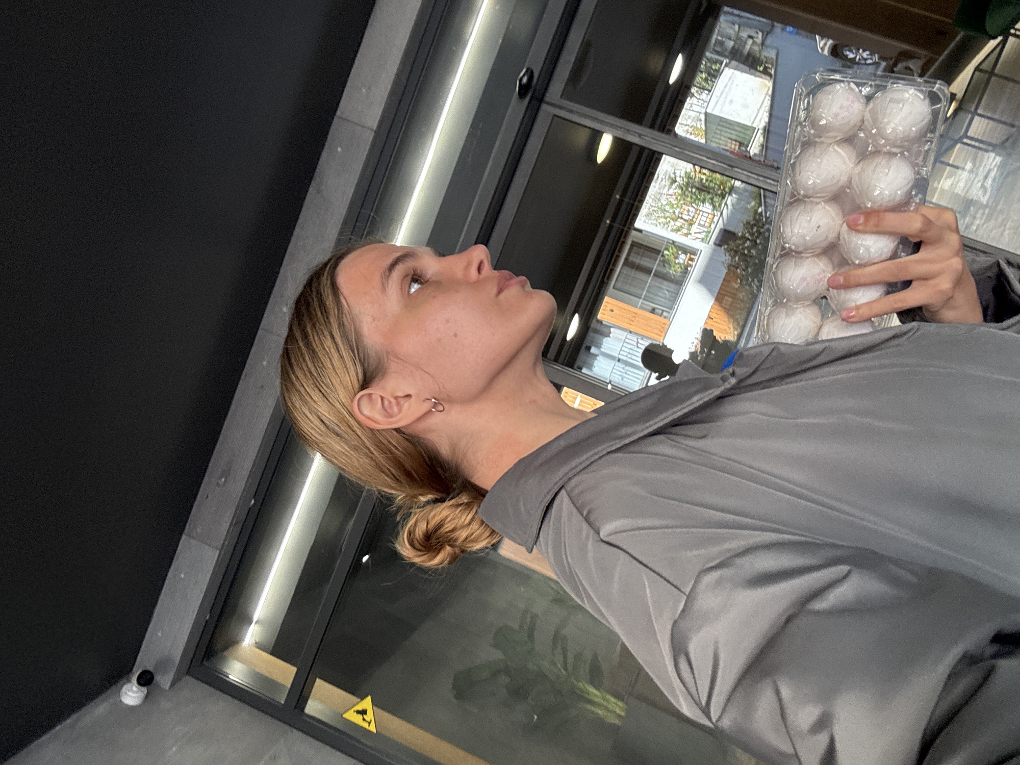 A medium shot, rotated to a Dutch angle, captures a young person, likely female, indoors during the daytime. She has blonde hair styled in a bun and wears a grey collared shirt or jacket. A silver hoop earring is visible in her left ear. Her head is tilted upwards, and she gazes towards the upper right of the frame (her right), with a neutral expression. In her right hand, she holds a clear plastic container housing twelve white, spherical items, which appear to be wrapped in a soft material and have faint pink markings.
The background reveals a contemporary interior with a dark grey wall on the left and expansive glass windows and doors on the right. Through these windows, blurred trees and other buildings suggest an outdoor urban or suburban setting. A small, black security camera is mounted on the wall in the lower left. A yellow triangular warning sign with a black symbol (possibly an electrical hazard) is visible on a window pane in the lower right. The bright lighting implies it is daytime. No discernible text is visible on the items or signs.