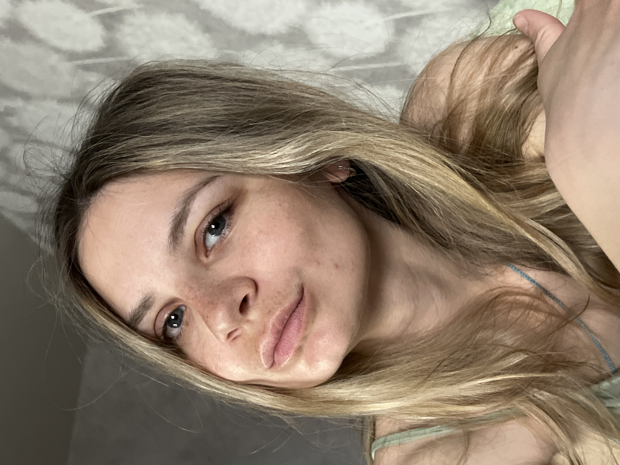A close-up, angled portrait of a fair-skinned woman with blonde hair. She has freckles and blue eyes. She is lying down, and her hair is spread around her head. She is wearing a light green tank top. The background appears to be a grey patterned surface, possibly a blanket or bedding. There is no information in the image to indicate it was taken in the city of Chaikovsky, Russia.