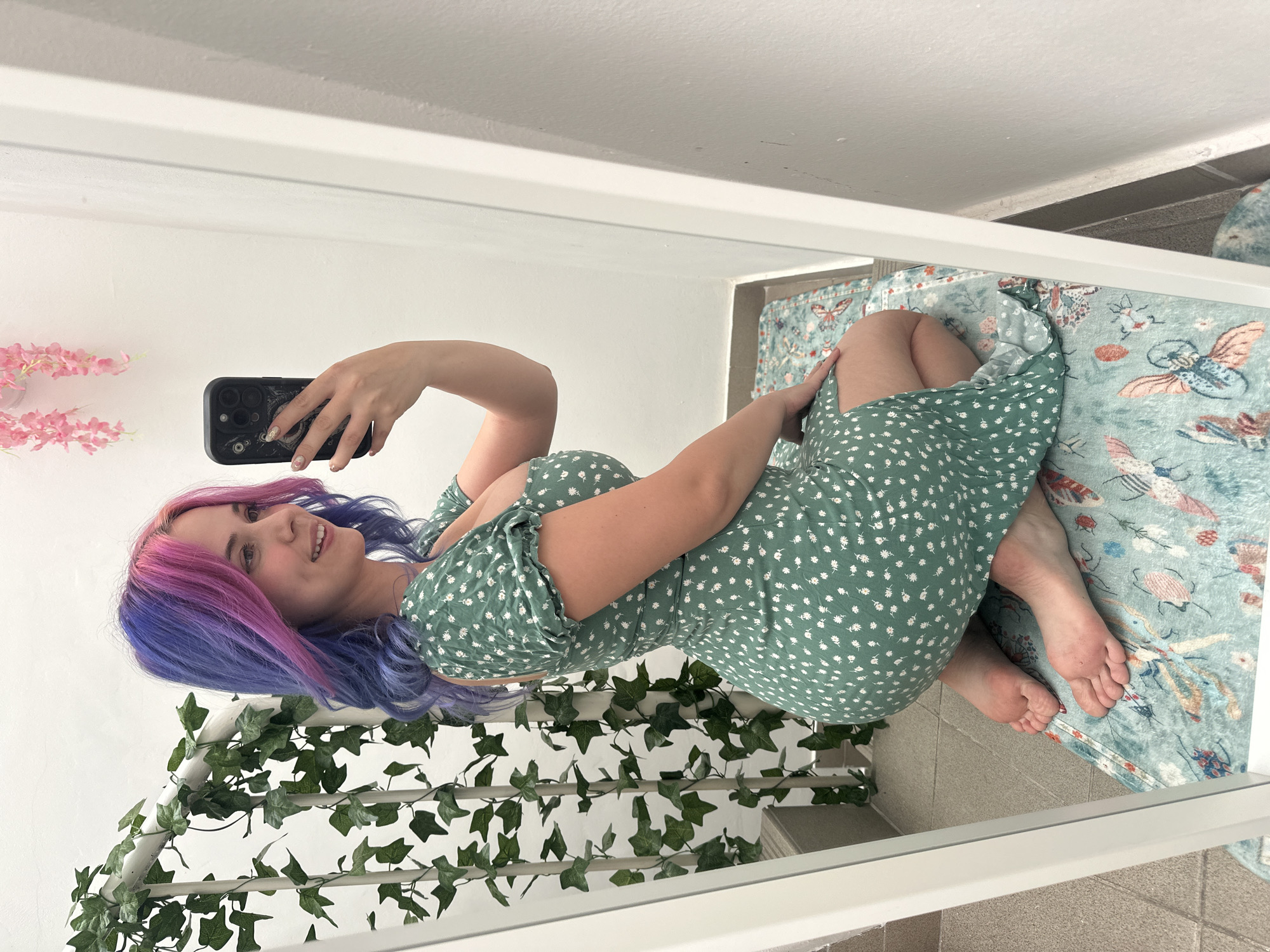 A young woman is captured in a mirror selfie, seen lying down and holding a black smartphone with her right hand to take the picture. She has striking two-toned hair, vibrant pink blending into purple, and is wearing a light green sleeveless dress adorned with small white floral patterns. A gentle smile graces her lips as she looks towards the phone's lens.
She appears to be resting on a bed or low platform, which is covered by a light blue-green blanket featuring various colorful insect and bird motifs. Her bare feet are visible on the blanket. Behind her, a white structure, possibly a bed frame or railing, is decorated with artificial green ivy vines. In the background to her left, against a white wall, are a few sprigs of artificial pink flowers. The floor visible in the reflection is tiled. The overall scene is indoors, presenting a casual and personal moment, likely within a residence in the City of Zagreb, Croatia. The image itself is rotated 90 degrees counter-clockwise. No visible text is present.