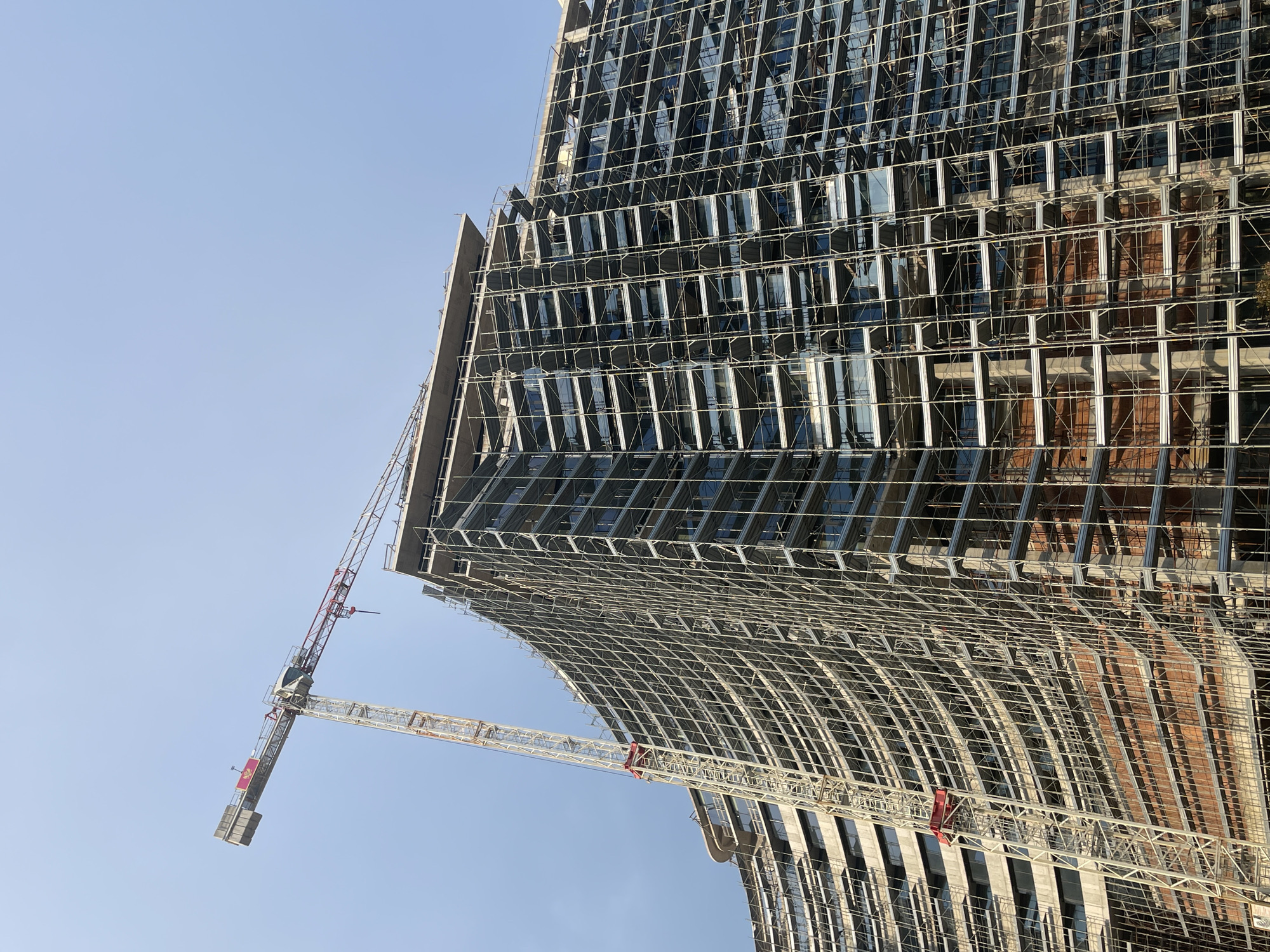 In Budva, Montenegro, a massive, partially constructed building is surrounded by extensive scaffolding. A large construction crane, painted white with red accents, extends diagonally across the sky from the left side of the image, its jib reaching over the top of the building. The building itself has a curved facade, with many floors visible, each covered in a dense network of metal scaffolding. Some windows are already installed, reflecting the clear blue sky. The lower sections of the building reveal exposed brickwork. The overall scene depicts a large-scale construction project in progress.