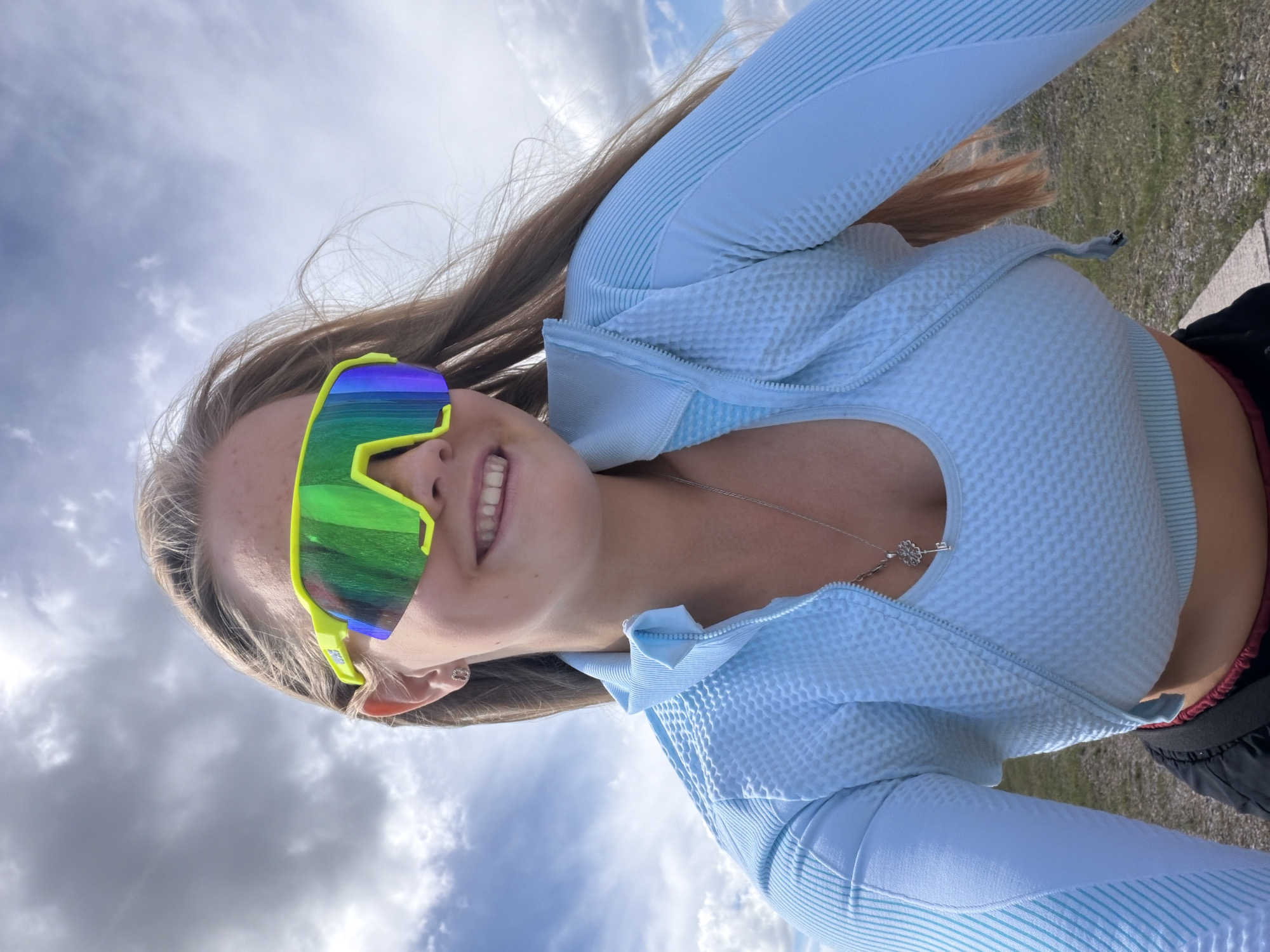 A young woman with long, light brown hair and fair skin is photographed outdoors, looking up and to her right with a smile. She is wearing bright yellow and green sunglasses with mirrored lenses, a light blue athletic top with a zipper, and a necklace with a key pendant. The background shows a cloudy sky and a blurred outdoor environment. The image appears to be taken in Chaikovsky, Russia, specifically near the Kama River, a popular area for outdoor recreation and scenic views in the city.
